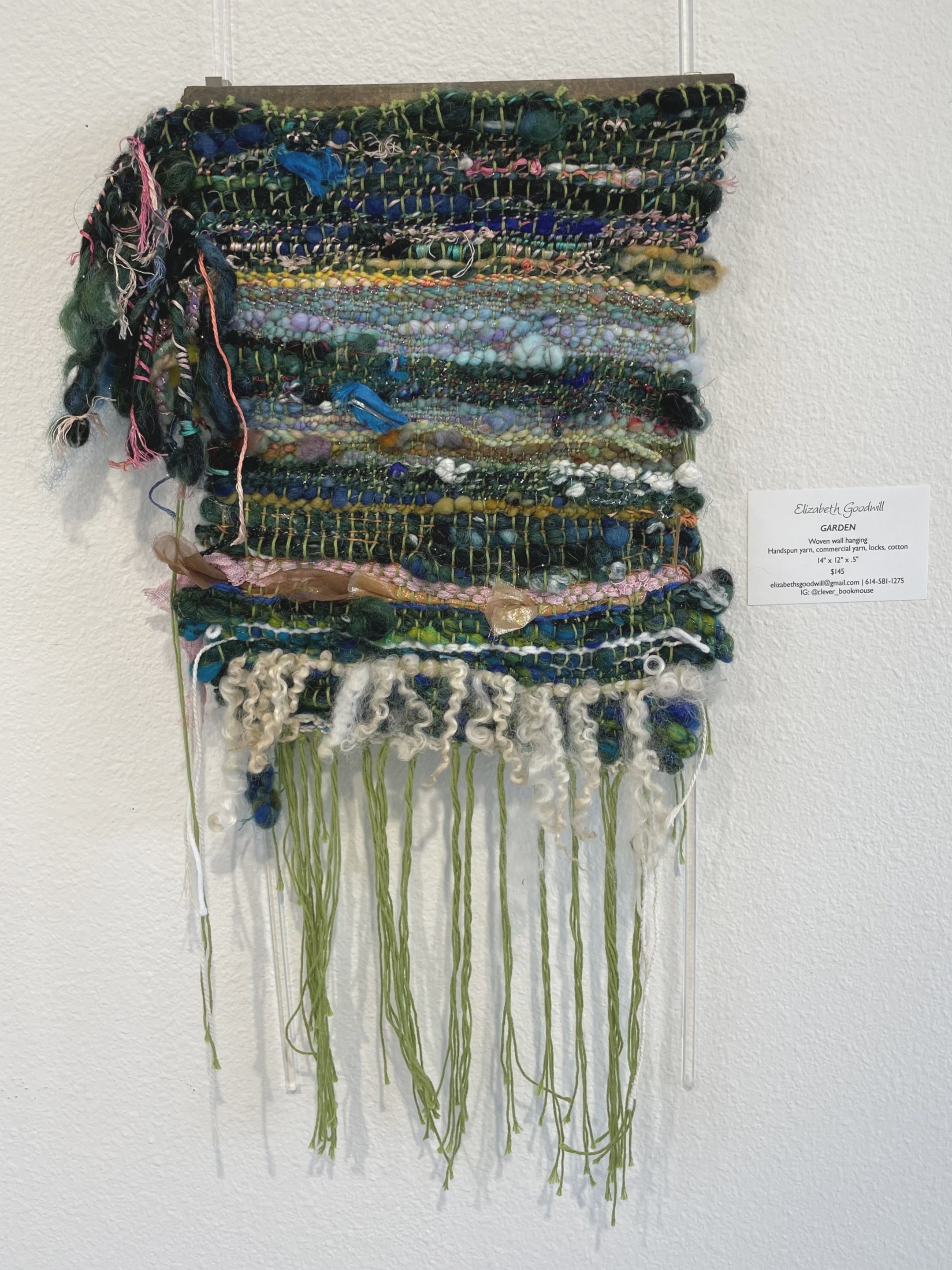 Mixed media wall hanging artwork with various fabric and yarn textures, hanging from a wooden rod, titled "Garden" by Elizabeth Goodwill.