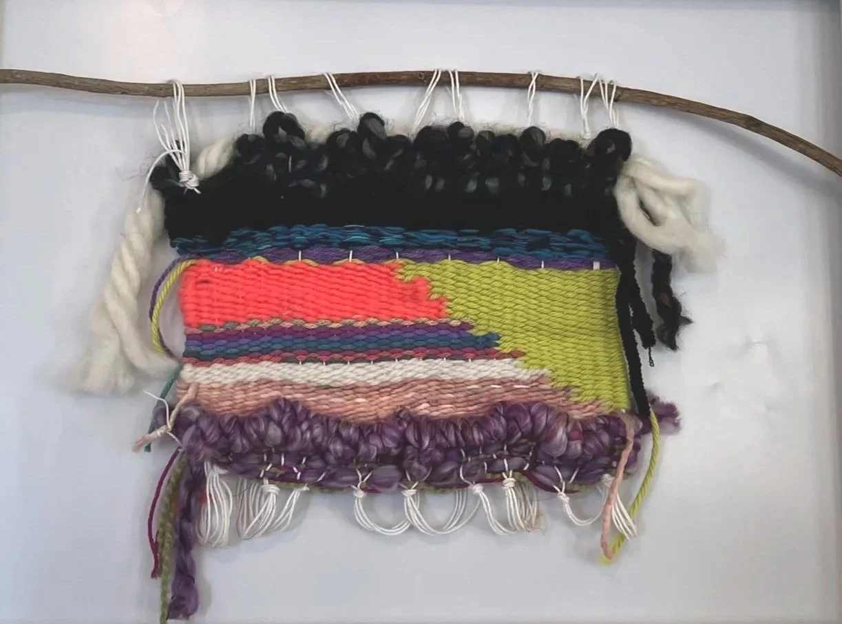 A colorful woven textile wall hanging with various yarn textures, colors, and patterns attached to a wooden stick.