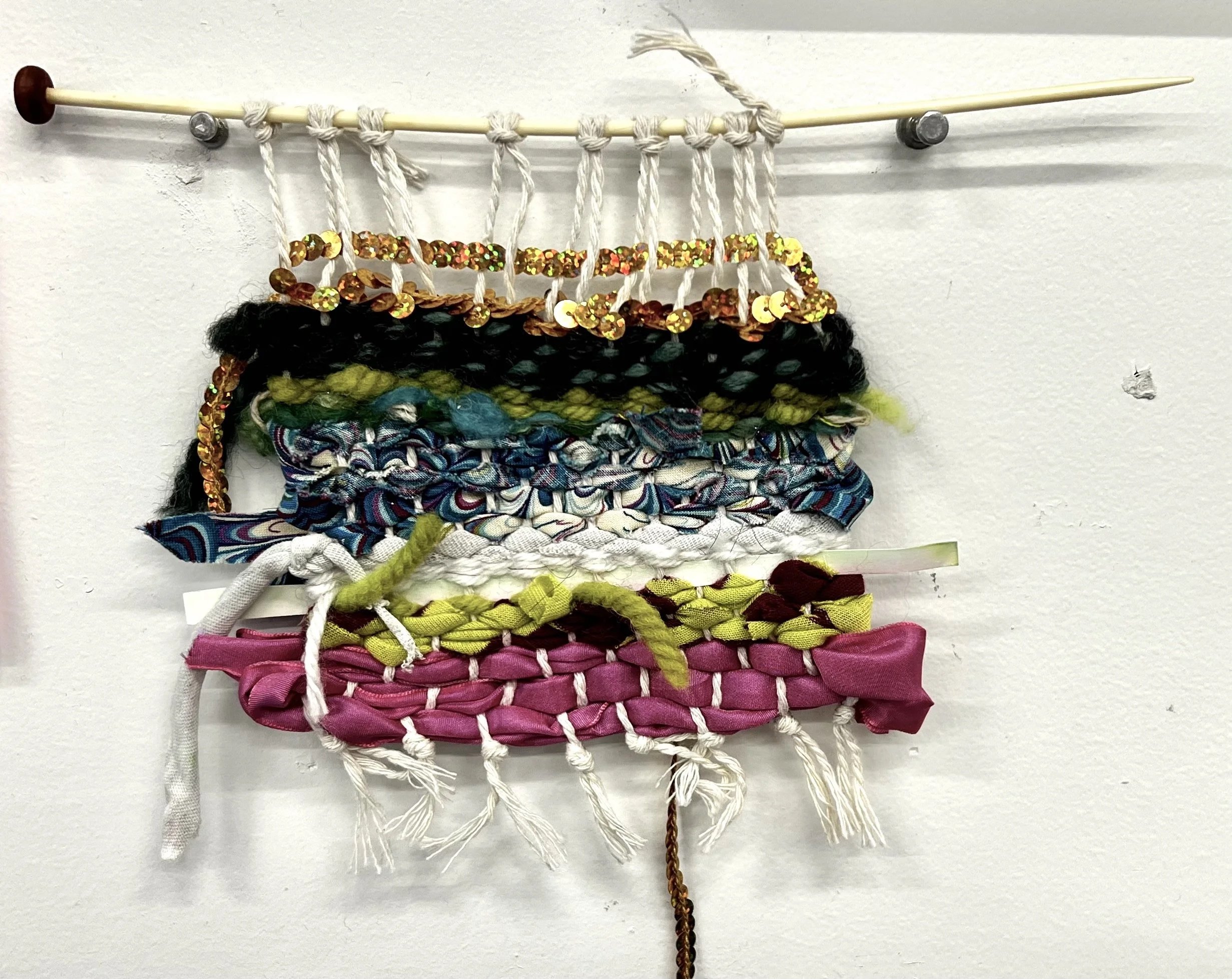 A wall hanging art piece made of various fabric strips, yarn, and sequins attached to a bar with strings.