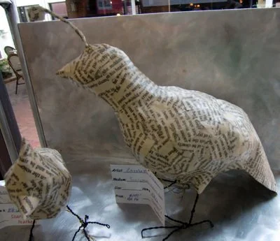 A bird sculpture made of pages of printed text, with wire legs and beak, on a metal surface in a display setting.