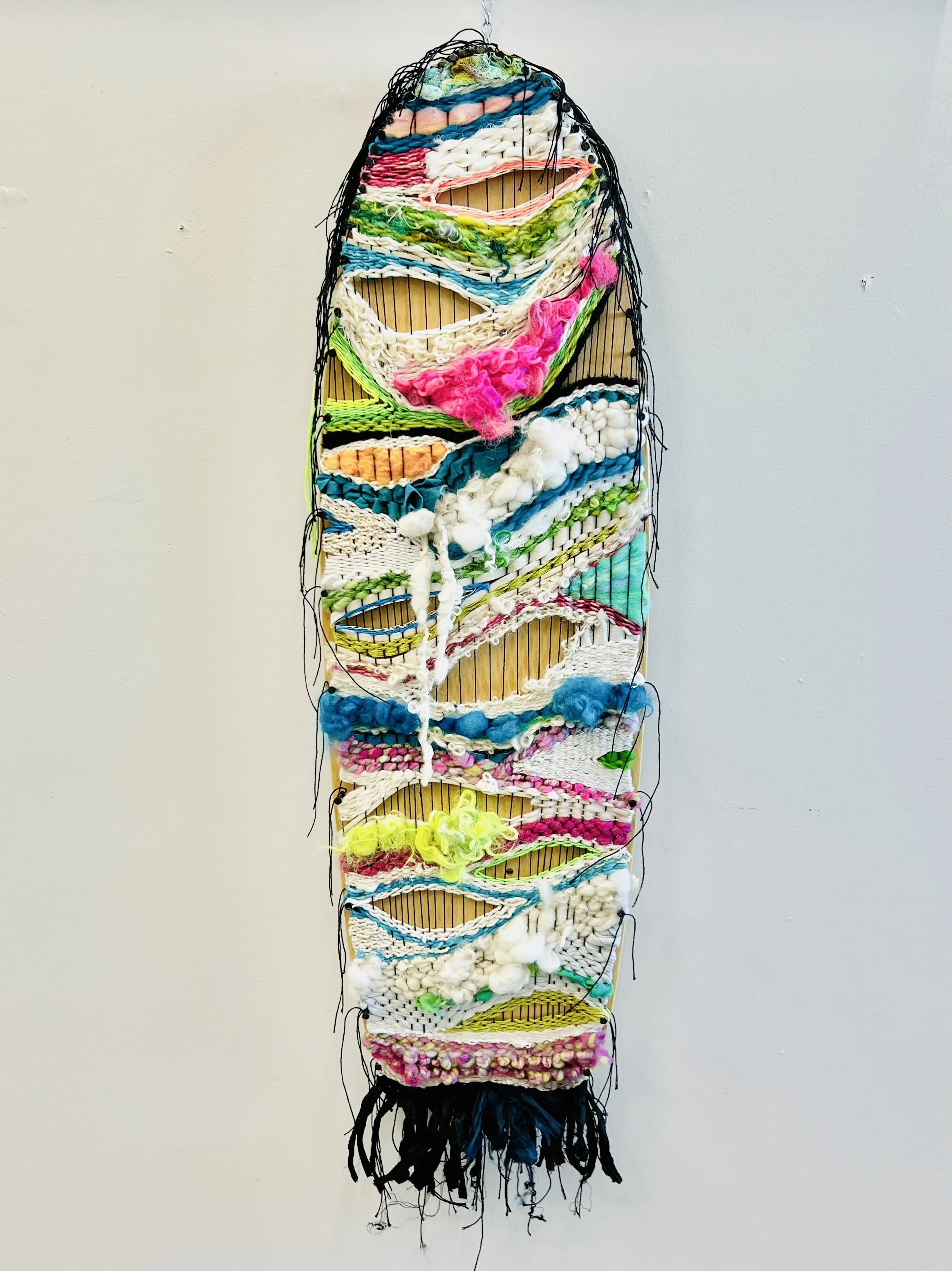 Colorful mixed-media textile wall art with various yarns, fabrics, and fibers arranged on a skateboard deck.
