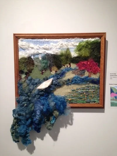 Mixed media collage of a landscape with mountains, trees, pink flowers, and water; textured fabric and yarn.
