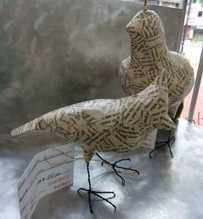 Two bird sculptures covered in newspaper print with wire legs and feet, displayed on a metal surface