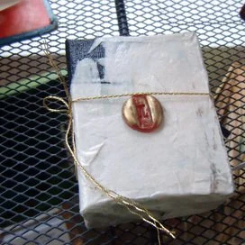 Wrapped gift box with a gold and red wax seal, tied with a gold string, on a black metal surface.