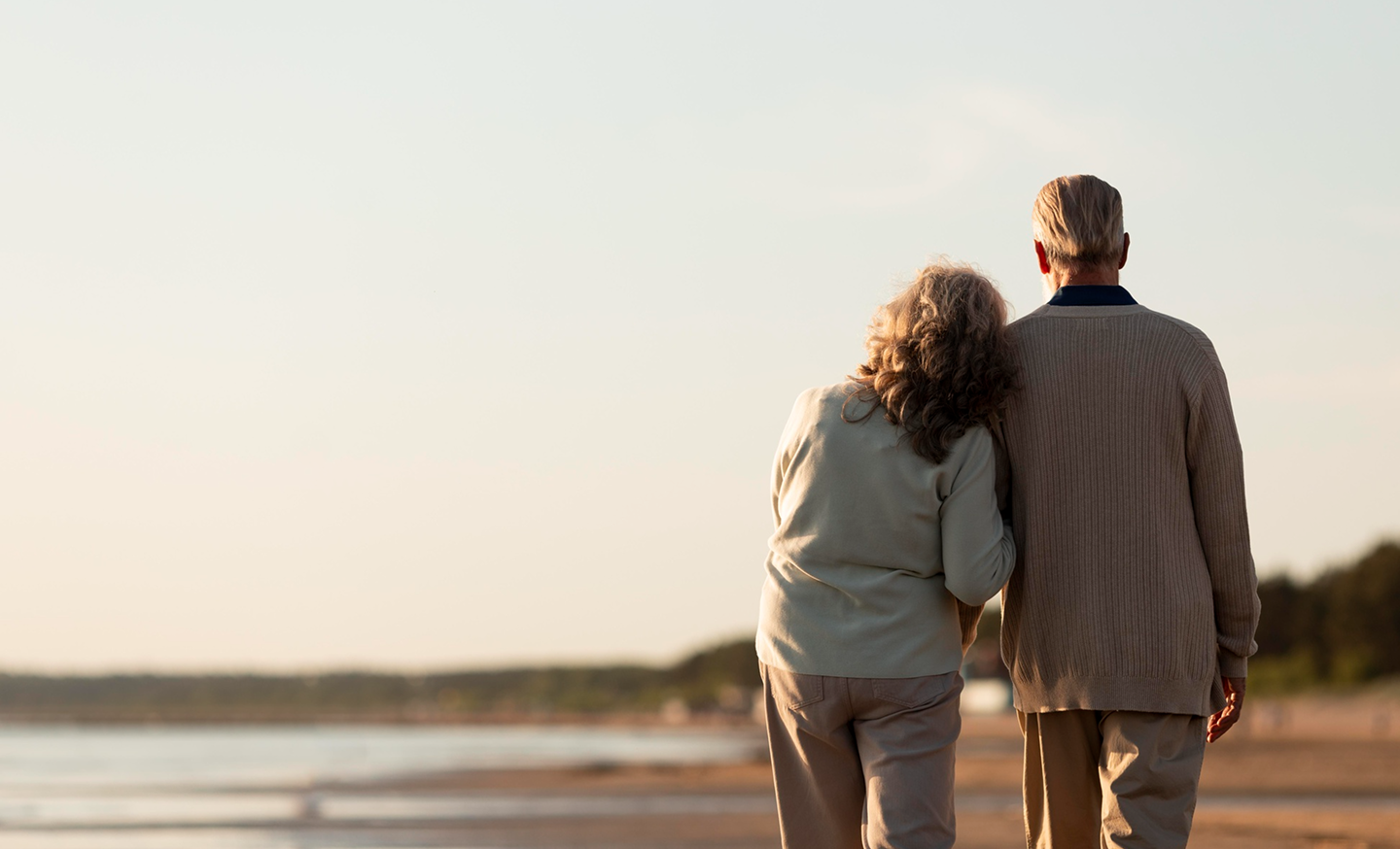 An elderly couple walking arm in arm along a beach at sunset.