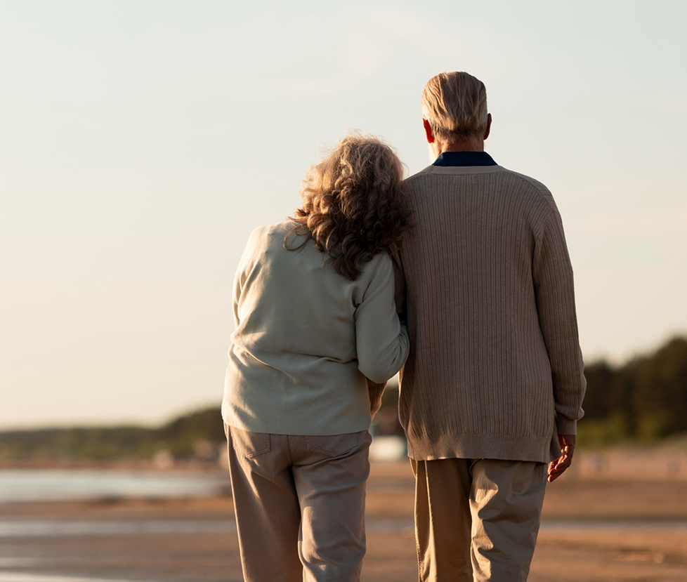 An elderly couple walking arm in arm on a beach at sunset.