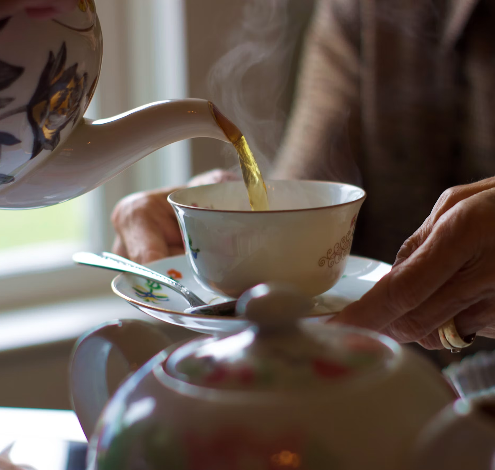 A person pouring hot tea from a floral teapot into a small tea cup. The person is holding the saucer with their other hand, and steam is rising from the tea.