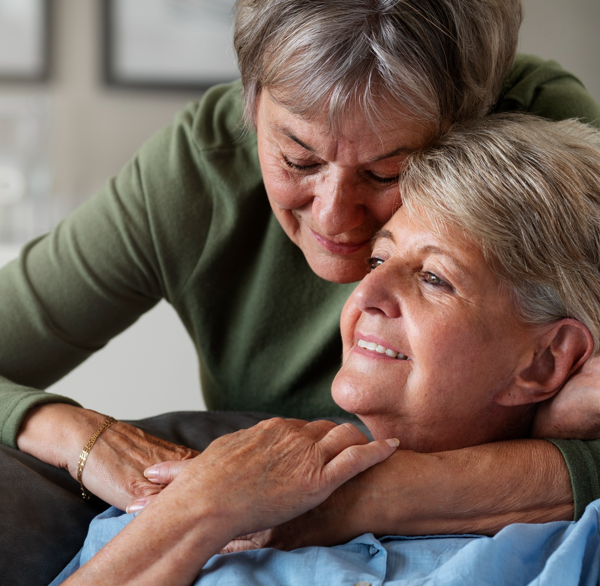 An elderly woman embracing an older person with short gray hair. The elderly woman has short gray hair, is smiling, and wears a green sweater. They are holding hands, and the scene suggests comfort and affection.