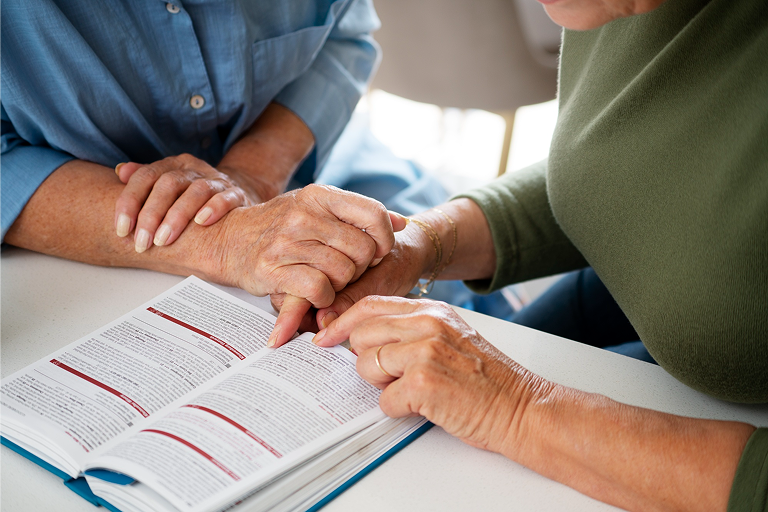 Two elderly women holding hands over an open book on a table.
