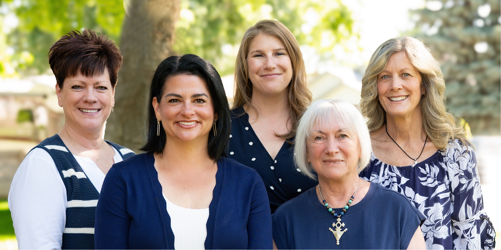Group of five women smiling outdoors with trees in the background.