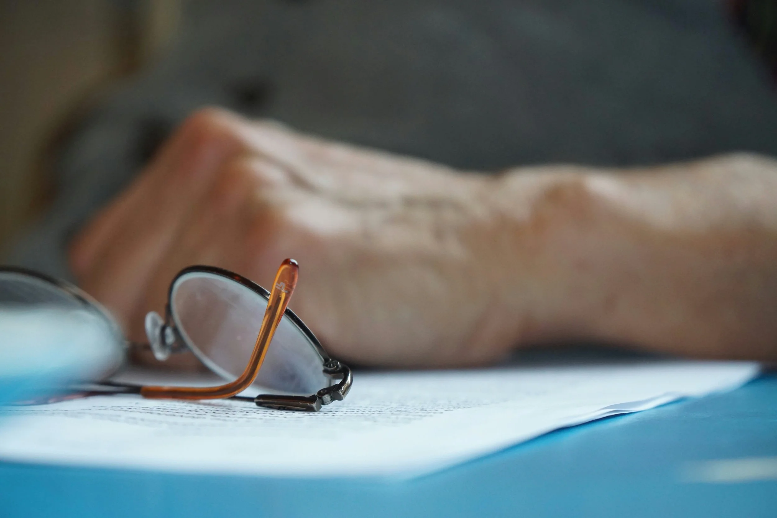 Close-up of eyeglasses resting on a surface, with a person's face and eye visible in the background.