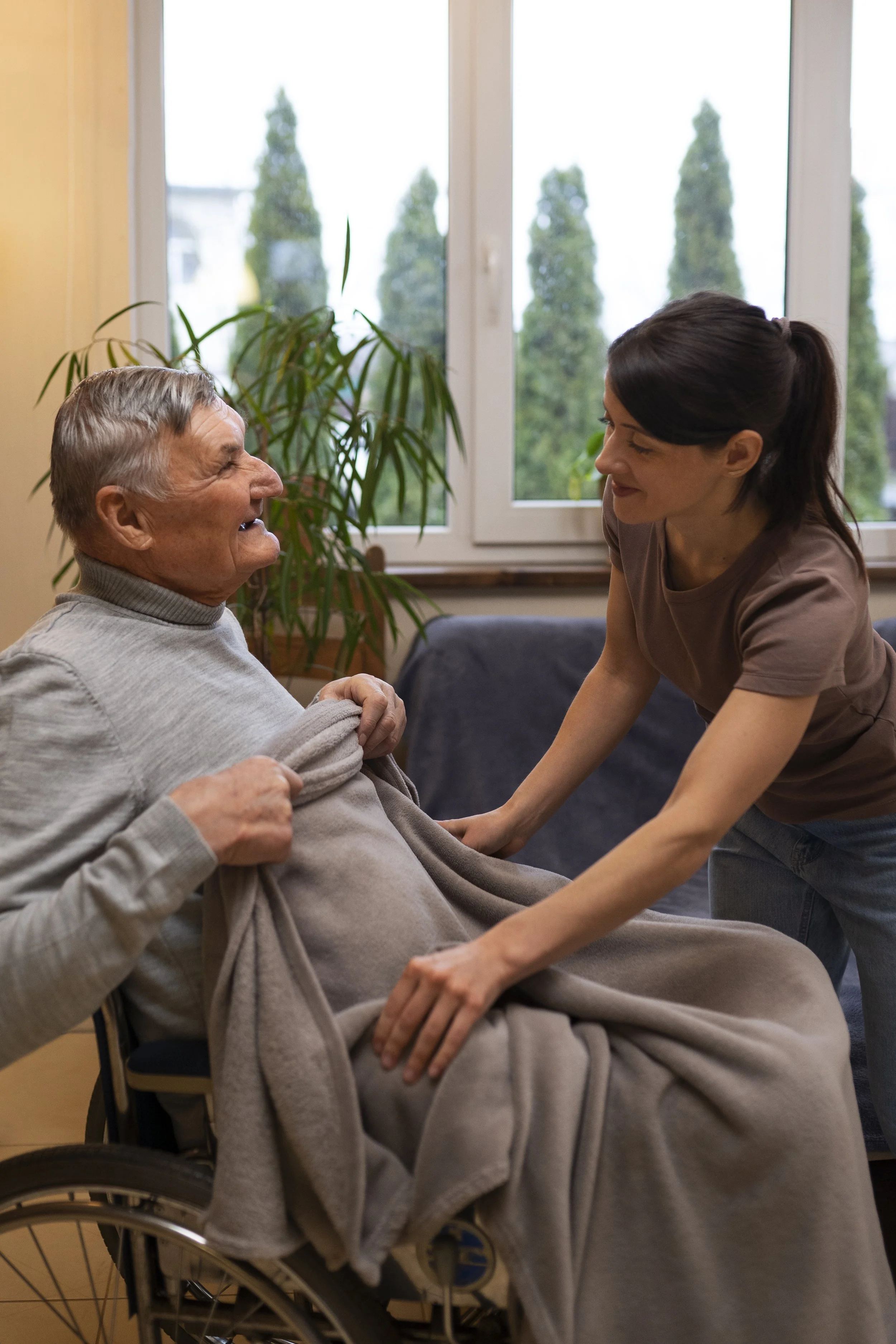 A young woman with dark hair tied back and a middle-aged senior man with gray hair using a wheelchair inside a room with large windows and a plant in the background. The woman appears to be assisting or caring for the man, holding his hands and smiling at him.