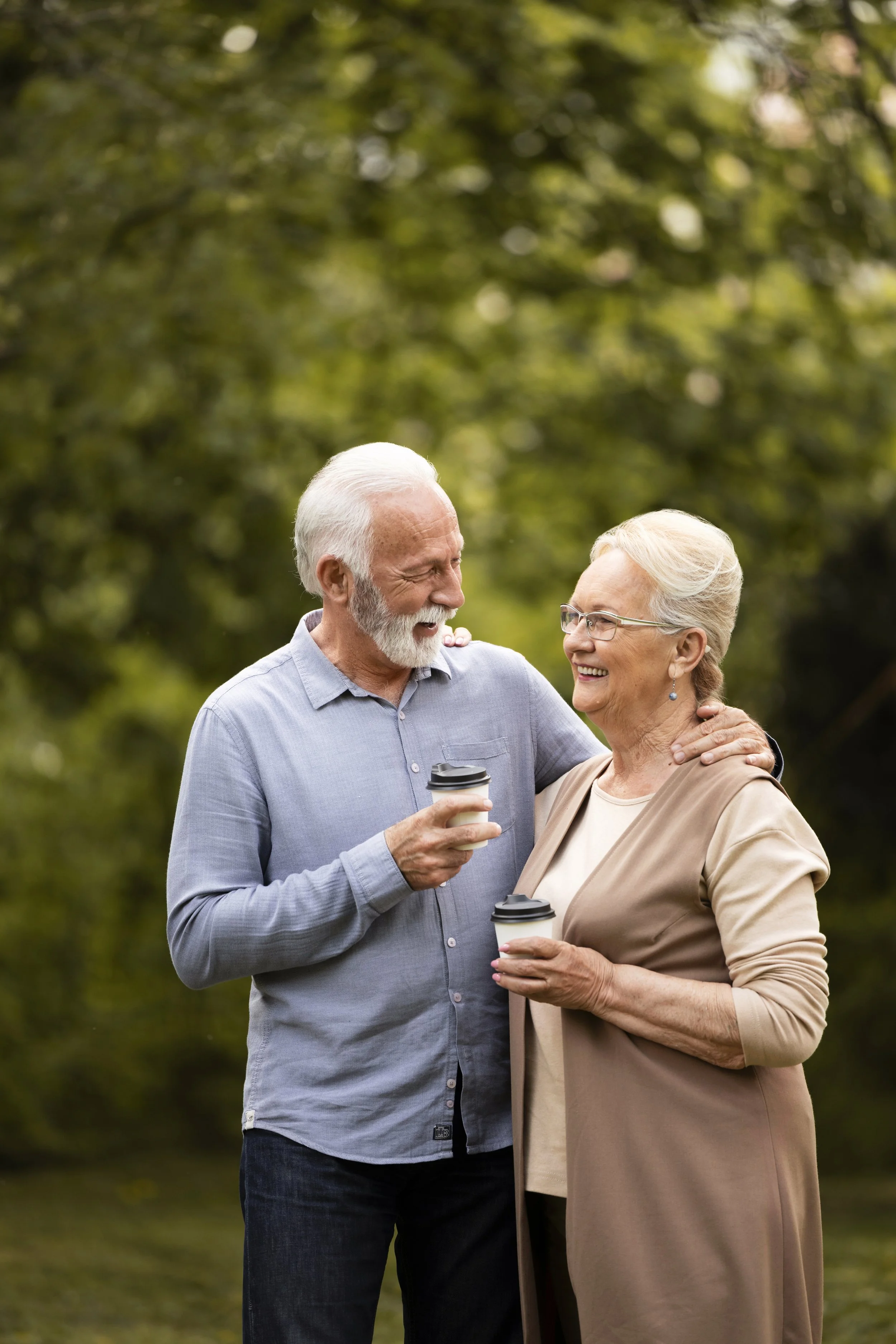 An elderly man and woman outdoors, smiling at each other and holding coffee cups, with blurred green trees in the background.