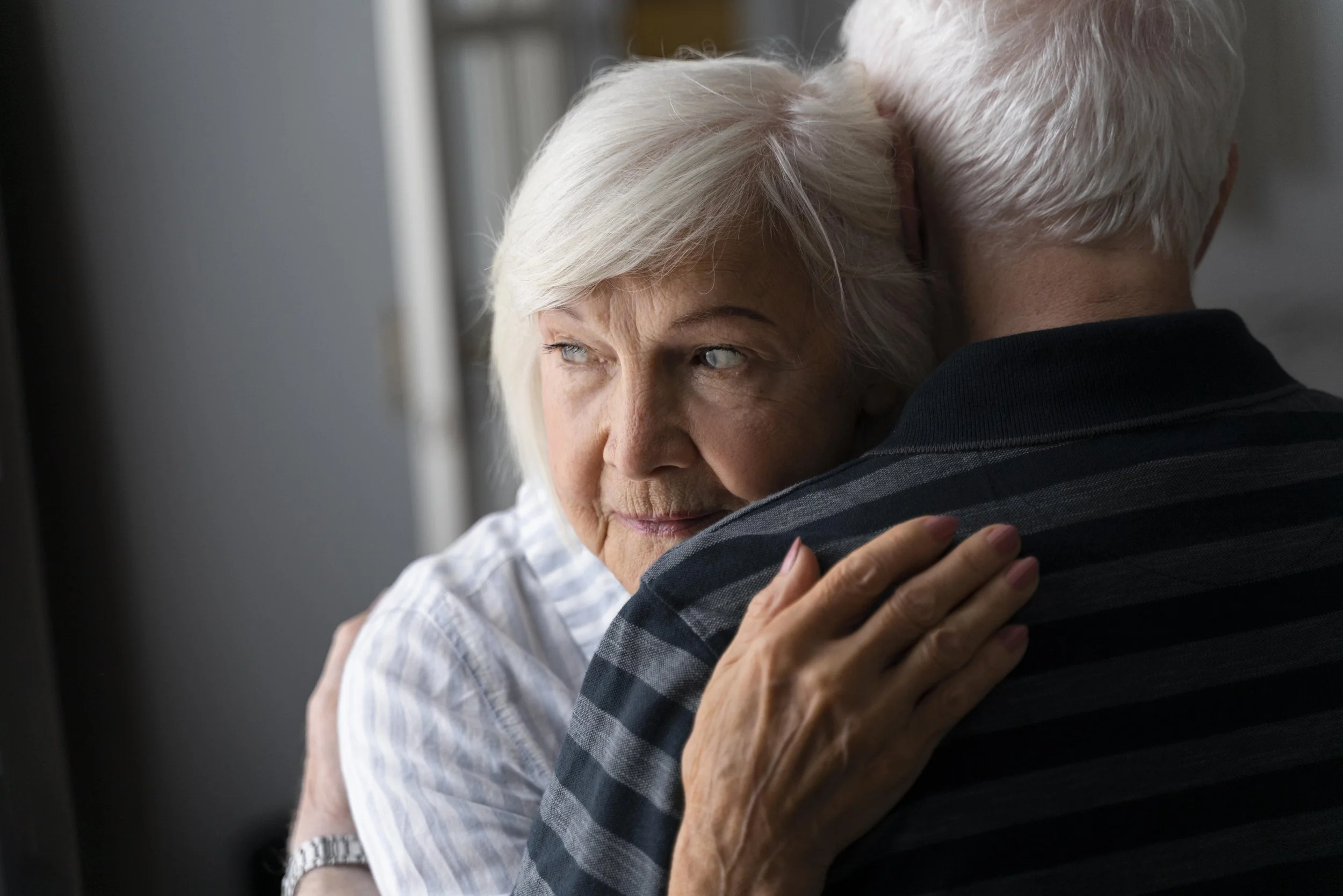 An elderly couple shares a warm embrace, with the woman resting her head on the man's shoulder and both smiling softly.