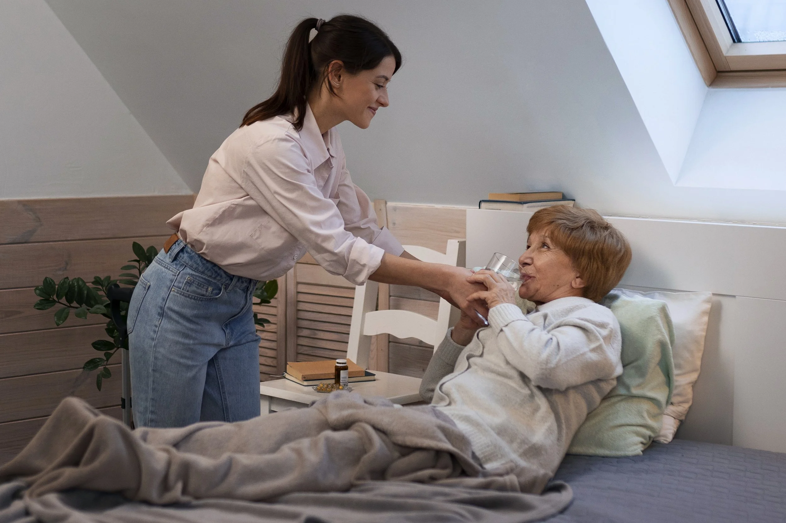 A young woman with dark hair in a ponytail is smiling and giving a glass of water to an elderly woman with short reddish hair, who is sitting in bed with a pillow behind her back, in a well-lit bedroom with skylight windows, wooden wall paneling, and some books on a white bedside table.