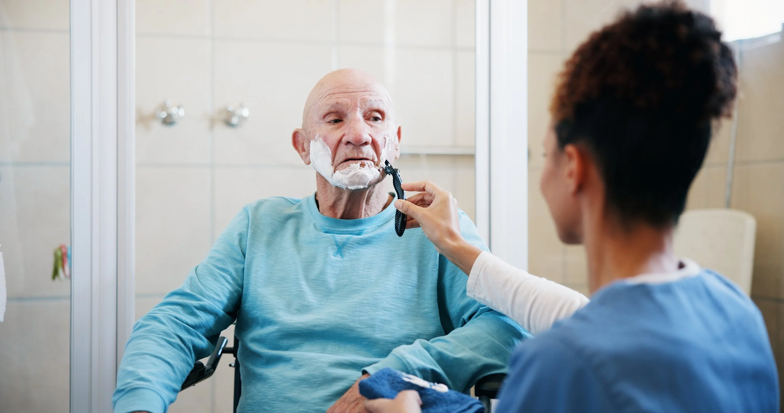 A young woman shaving an elderly man with a beard trimmer in a bathroom.