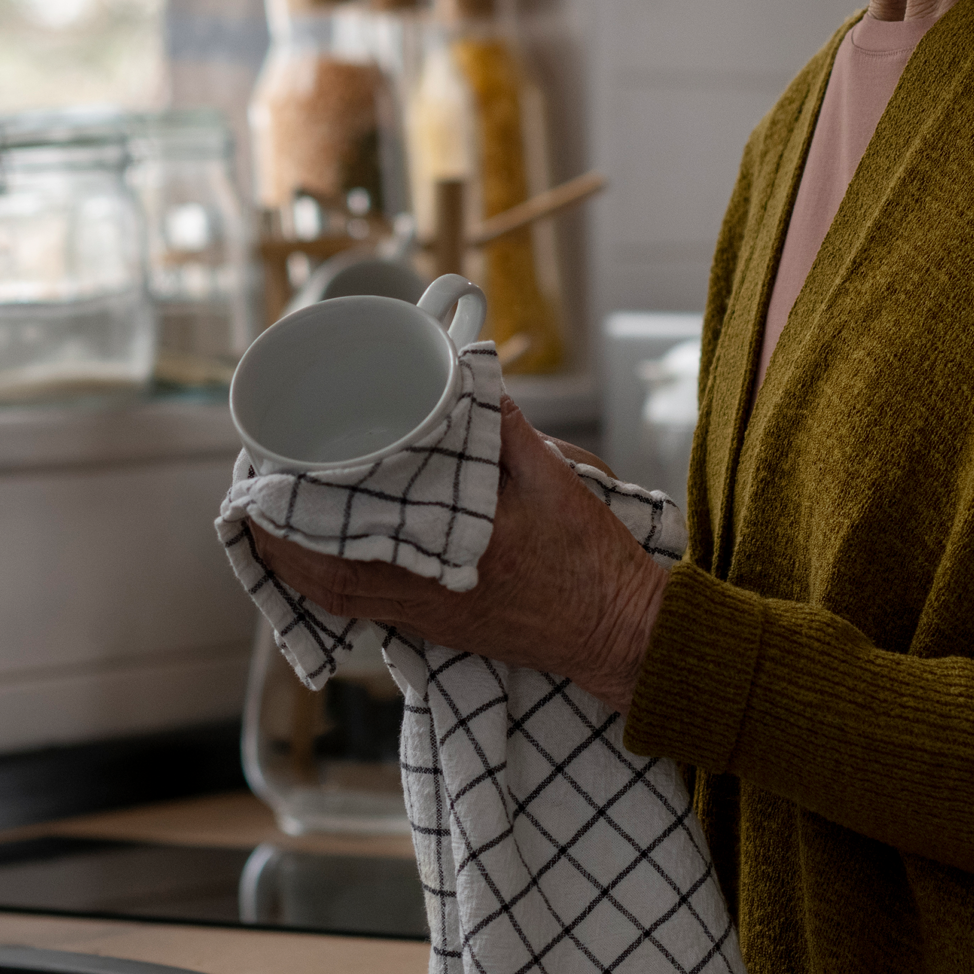 Person in mustard yellow sweater holding a white coffee mug wrapped in a checkered cloth in a kitchen.