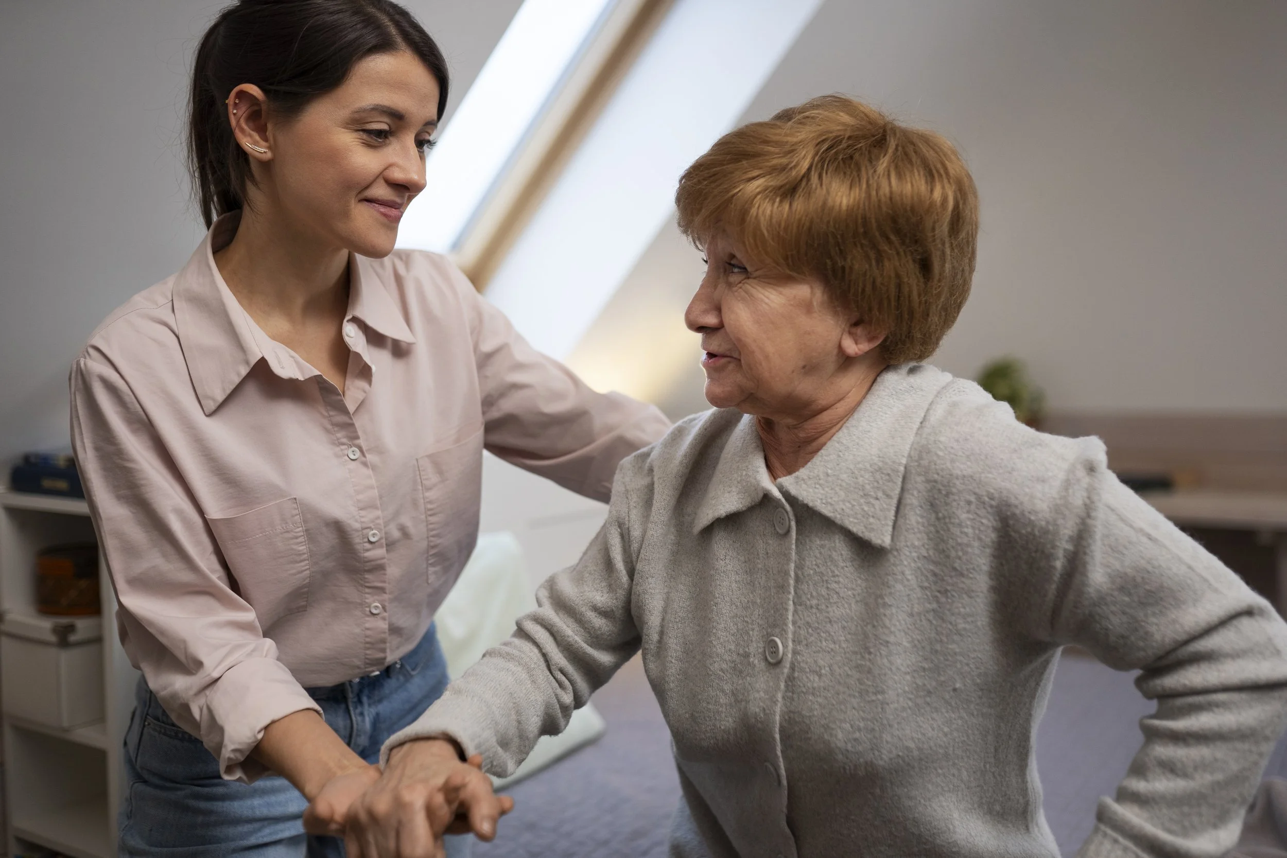 A young woman helping an elderly woman stand up, in a bright room with a sloped window in the background.