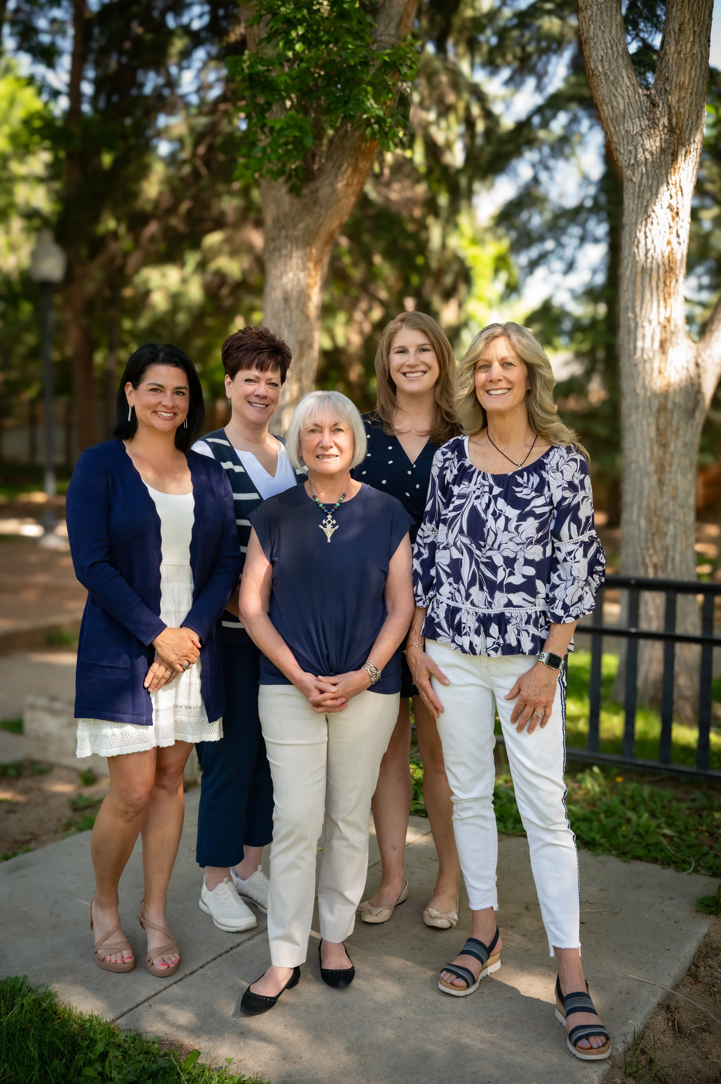 Group of five women standing outdoors on a sidewalk with trees in the background, smiling at the camera.