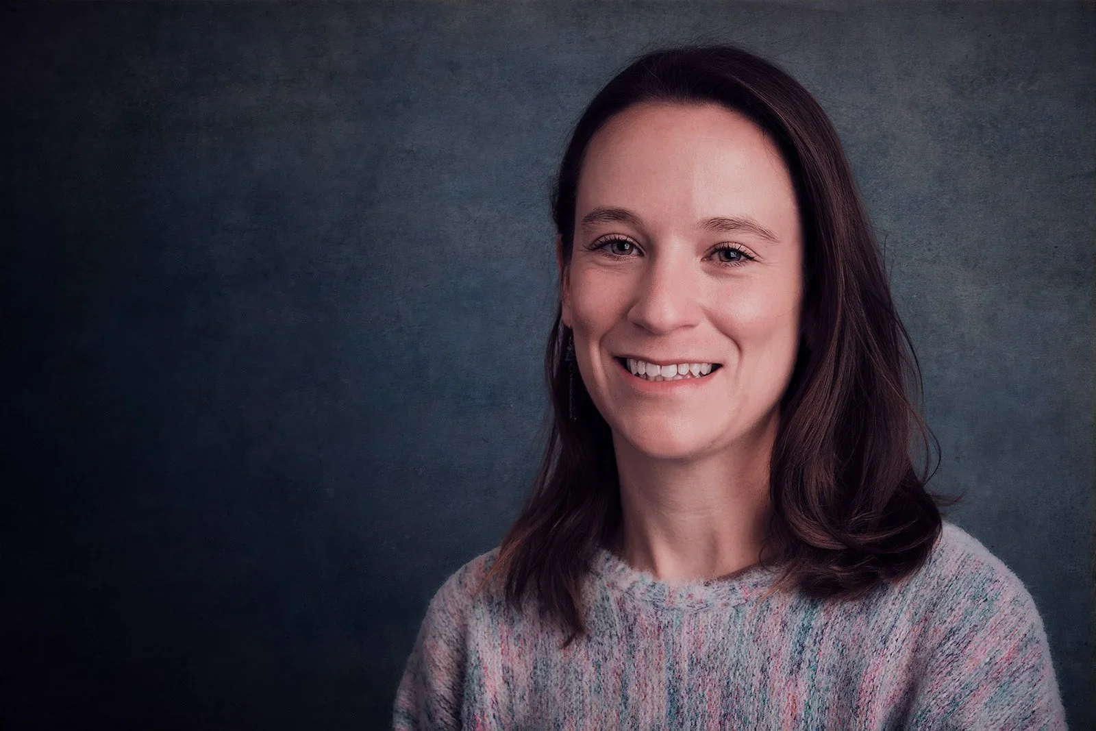 Smiling woman with dark brown hair and blue eyes wearing a multicolored sweater against a dark, textured background.