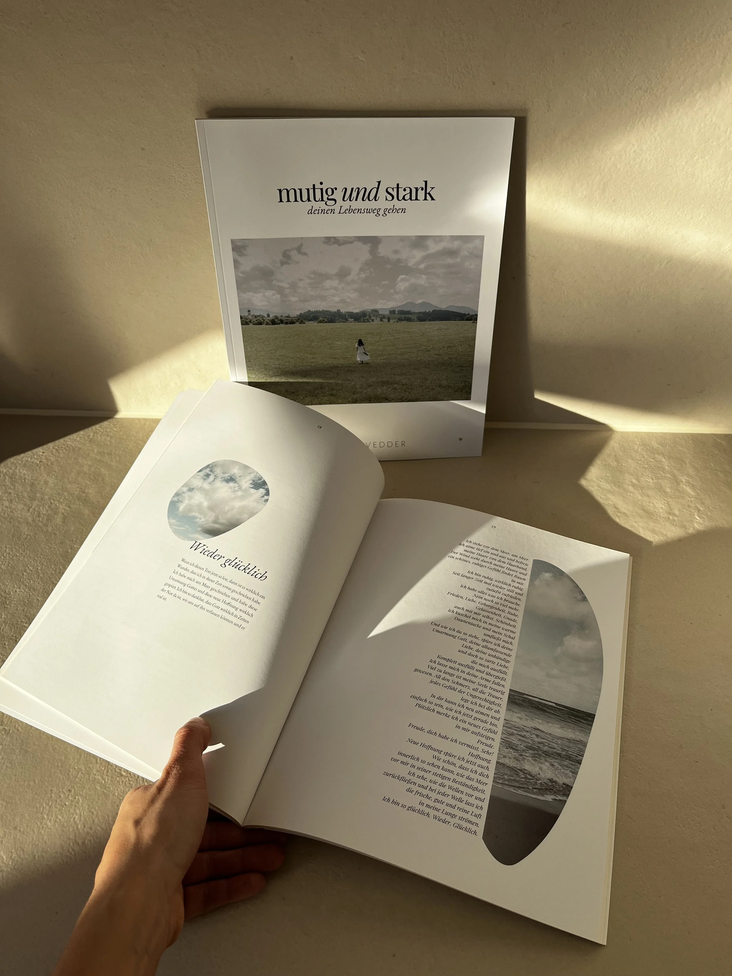 Offenes Buch mit Text und Bildern, im Hintergrund ein Stand mit Buch, auf einem Tisch im Sonnenlicht.