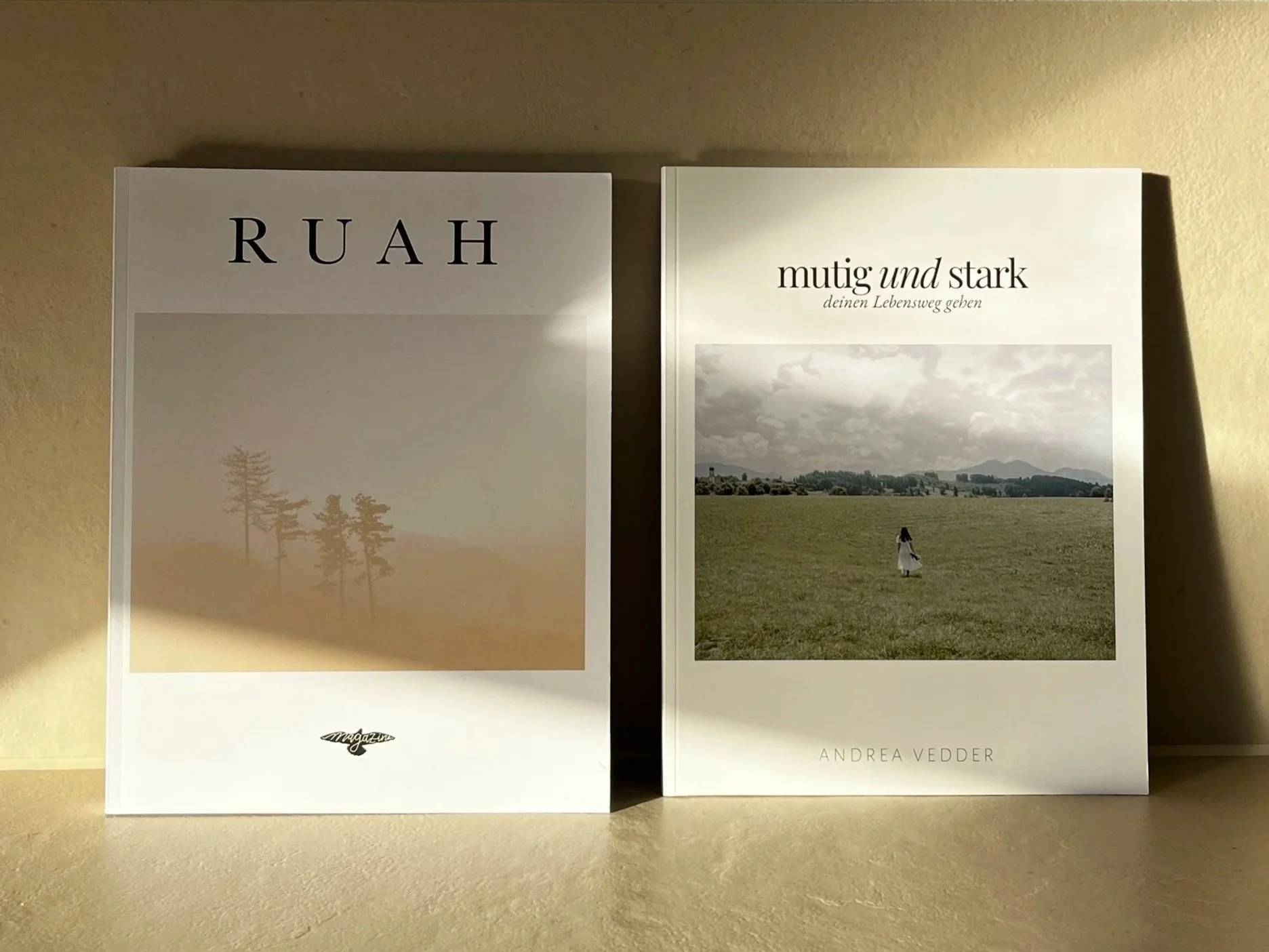 Zwei Bücher stehen auf einer Oberfläche. Das linke Buch trägt den Titel "RUH" mit einem Bild von dünnen Baumlinien in einer ländlichen Landschaft. Das rechte Buch trägt den Titel "mutig und stark" mit einem Bild eines kleinen Mädchens in einem weißen