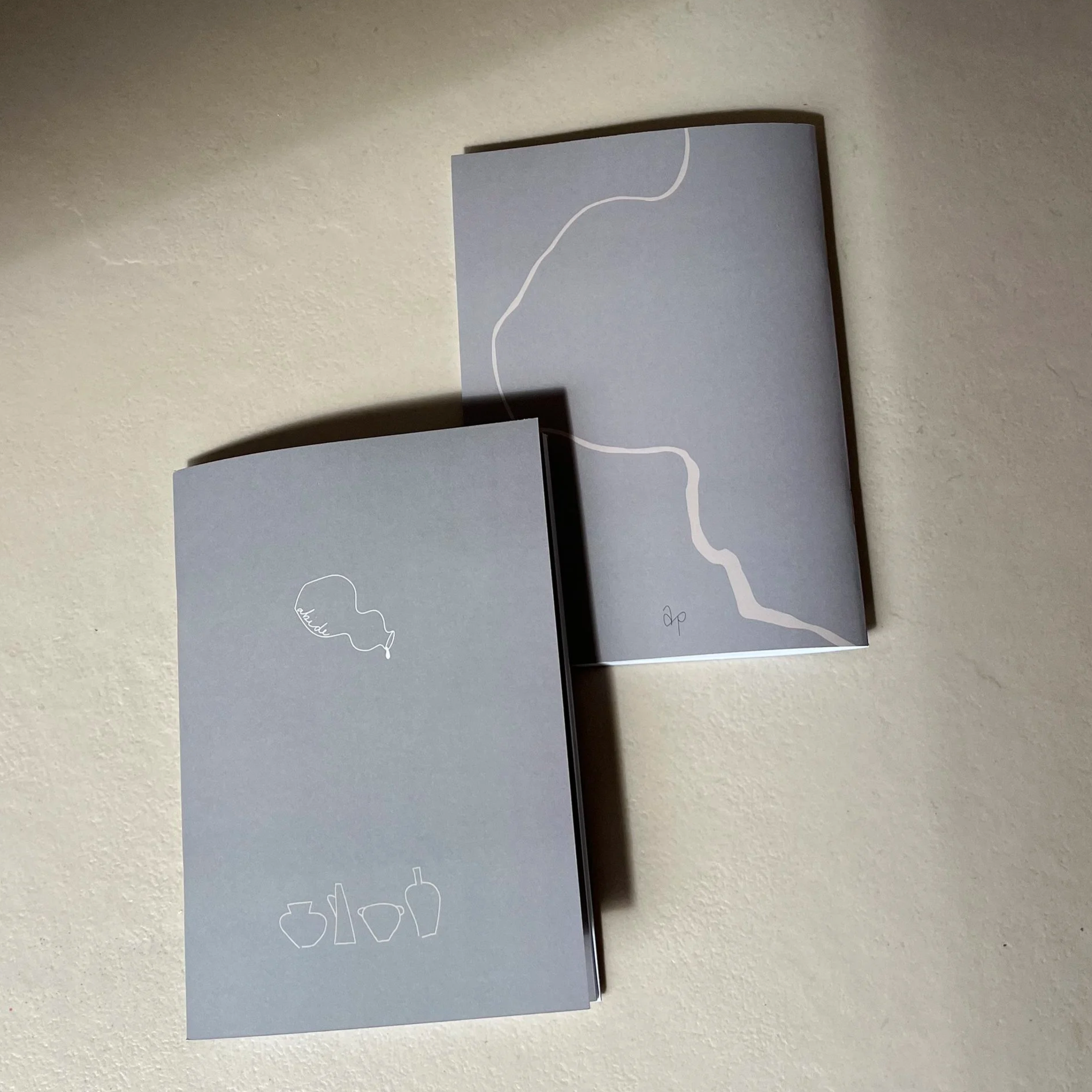 Zwei Bücher oder Kataloge mit hellgrauem Einband, jeweils mit minimalistischen weißen Linienzeichnungen von Geschirr und einer Linie, die eine Landschaft oder einen Fluss darstellt, liegen auf einer weißen Oberfläche.