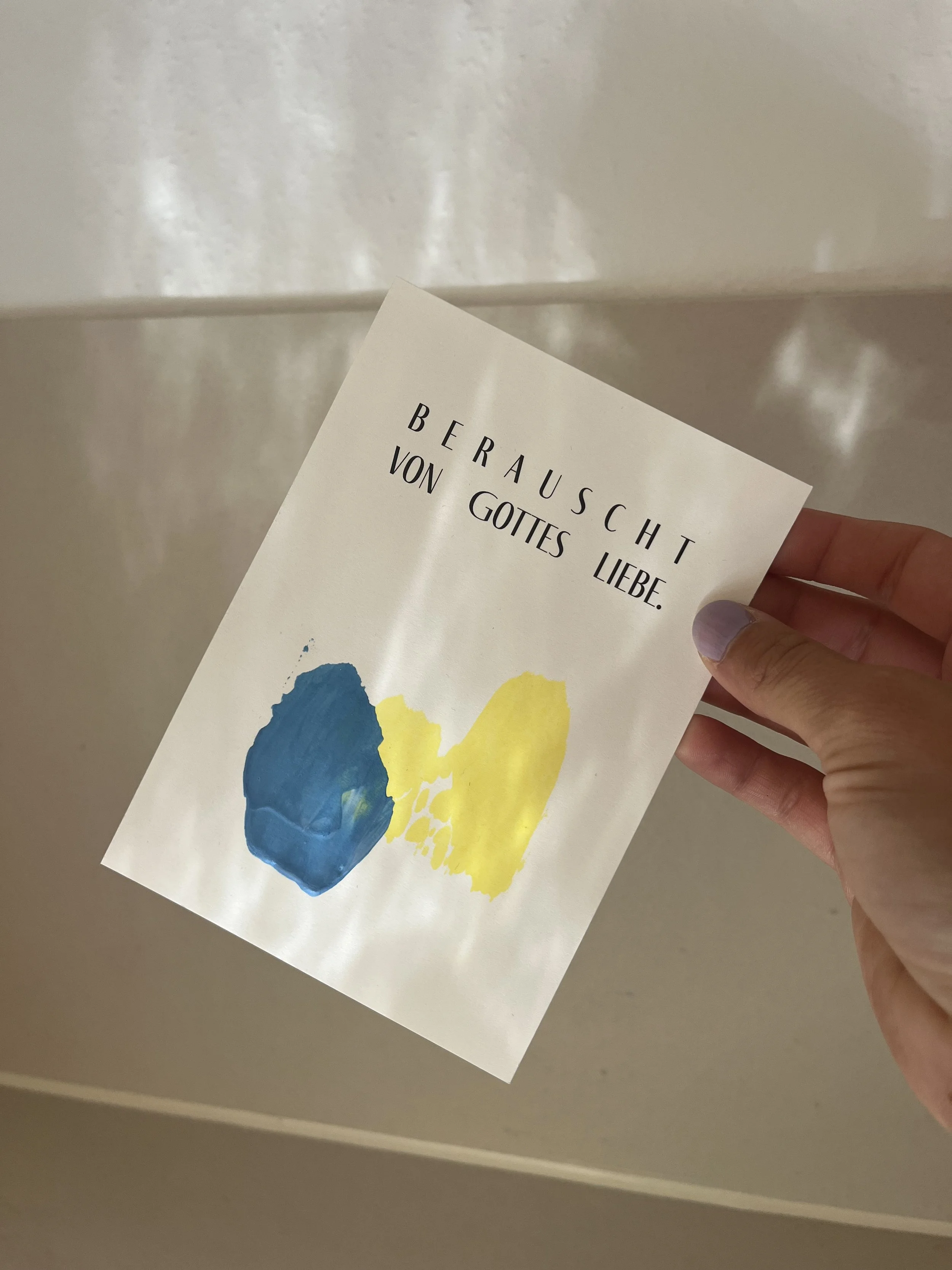 Eine Hand hält eine Karte mit der Aufschrift 'Berührung von Gottes Liebe' und zwei Malerei, eine blaue und eine gelbe, auf weißem Hintergrund.