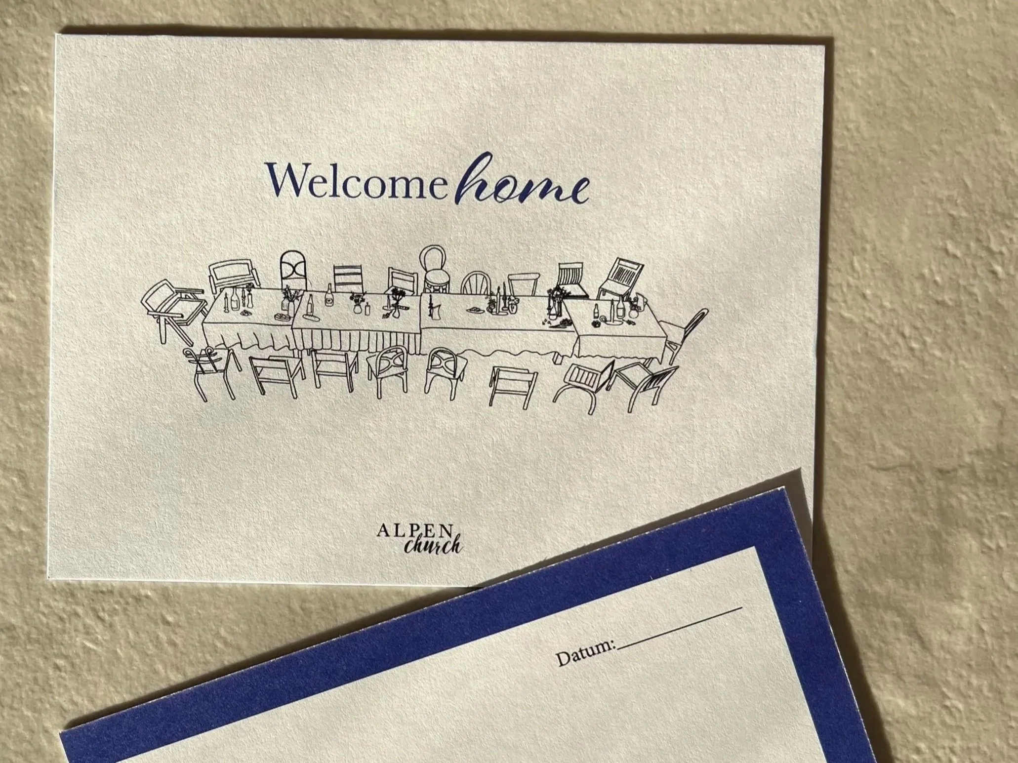 Willkommenskarte mit einem zeichnerischen Bild eines Tisches mit Stühlen und Dekorationen, darunter die Worte "Welcome home" und das Logo "Alpen Church".