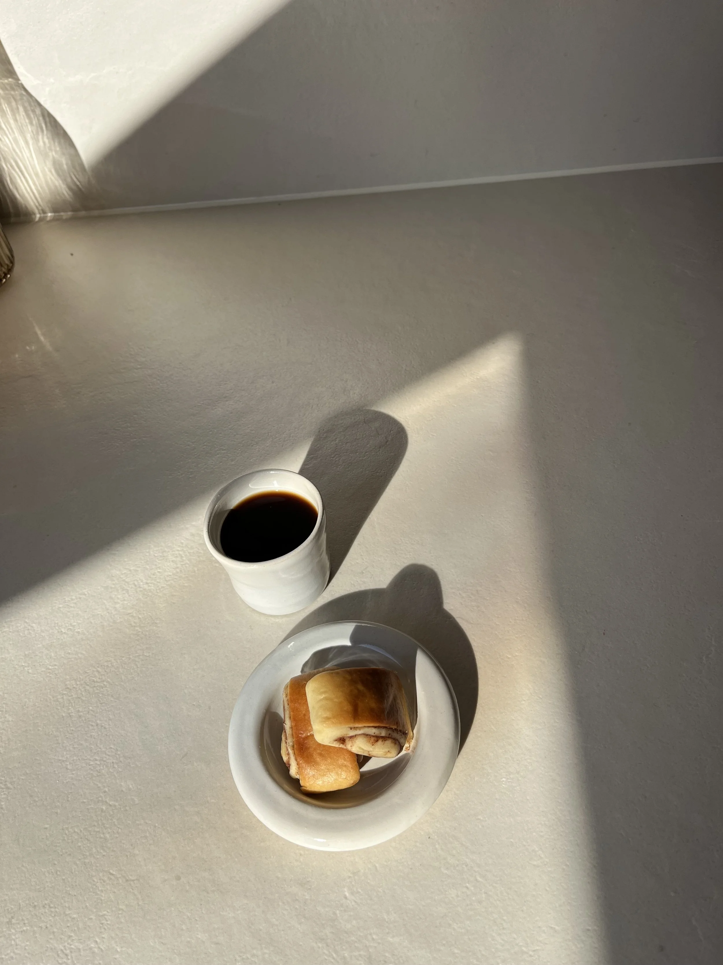 Tasse Kaffee und ein Teller mit zwei Gebäckstücken auf einem hellen Tisch, mit Sonnenlicht und Schatten.
