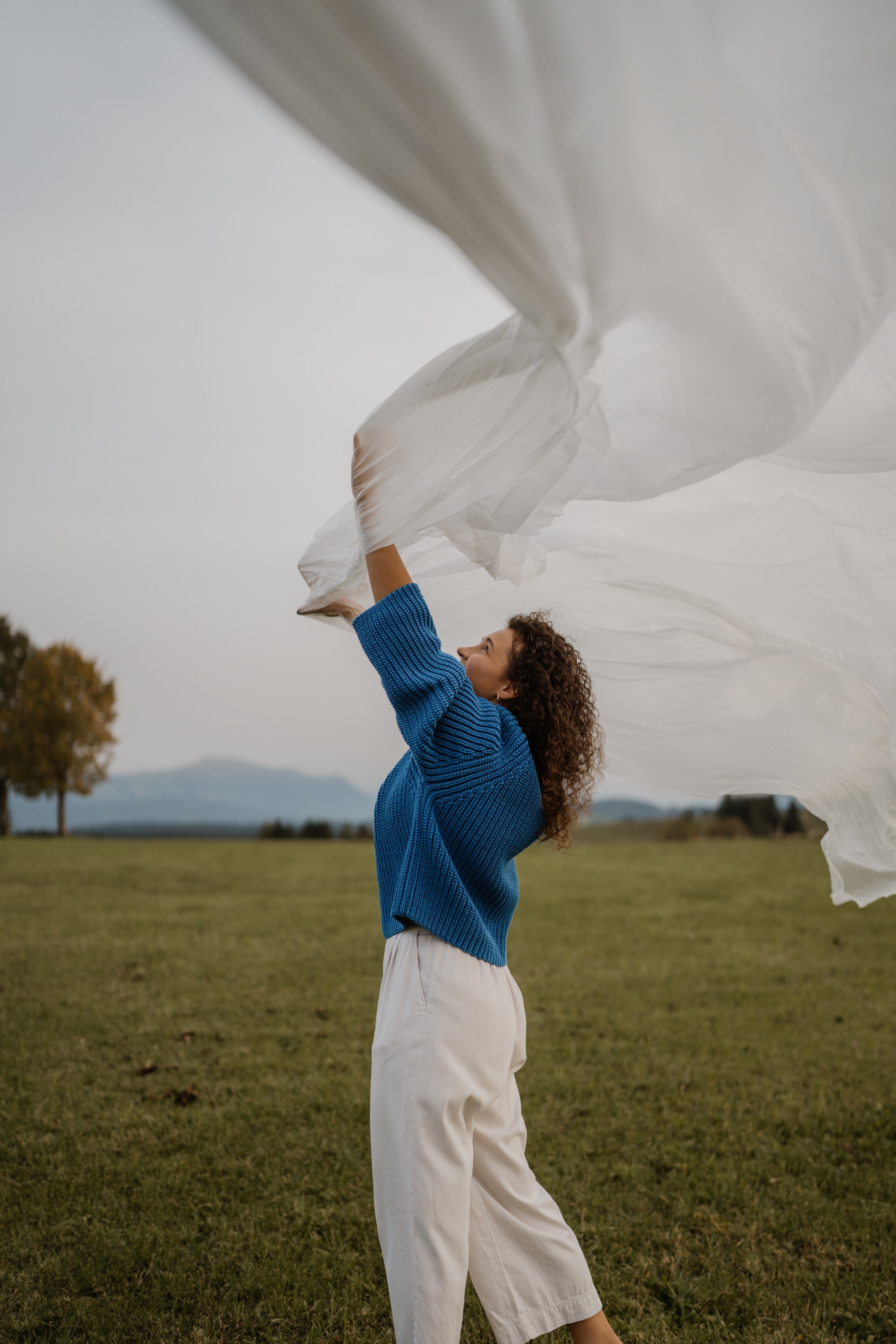 Frau im blauen Pullover und weißen Hosen, die auf einer Wiese einen weißen Stoff in die Luft wirft.