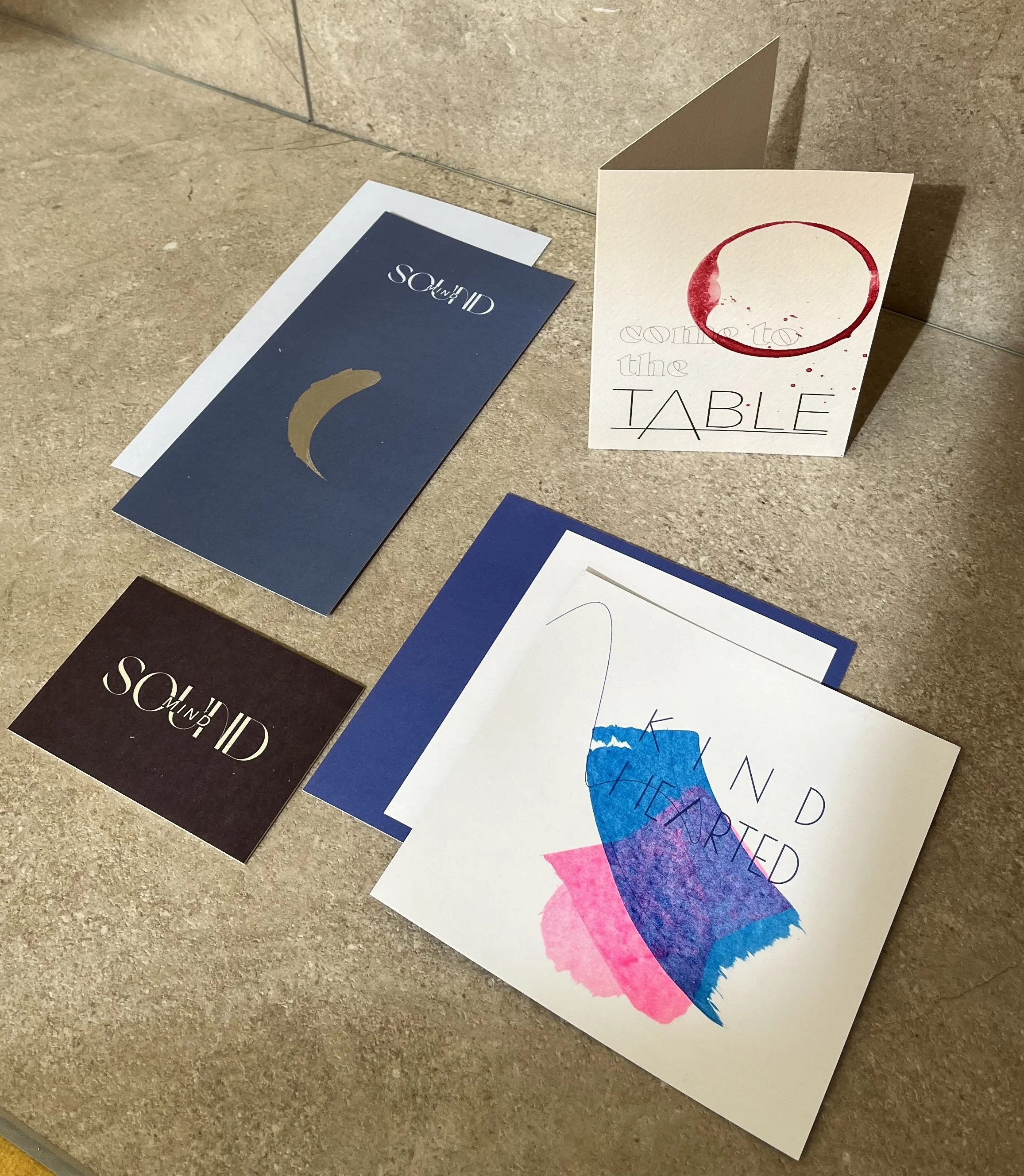 Mehrere Karten auf einem Boden, eine mit einem roten Kaffeefleck, eine mit buntem Wasserfarbenkussel, andere mit Texten und Designs.