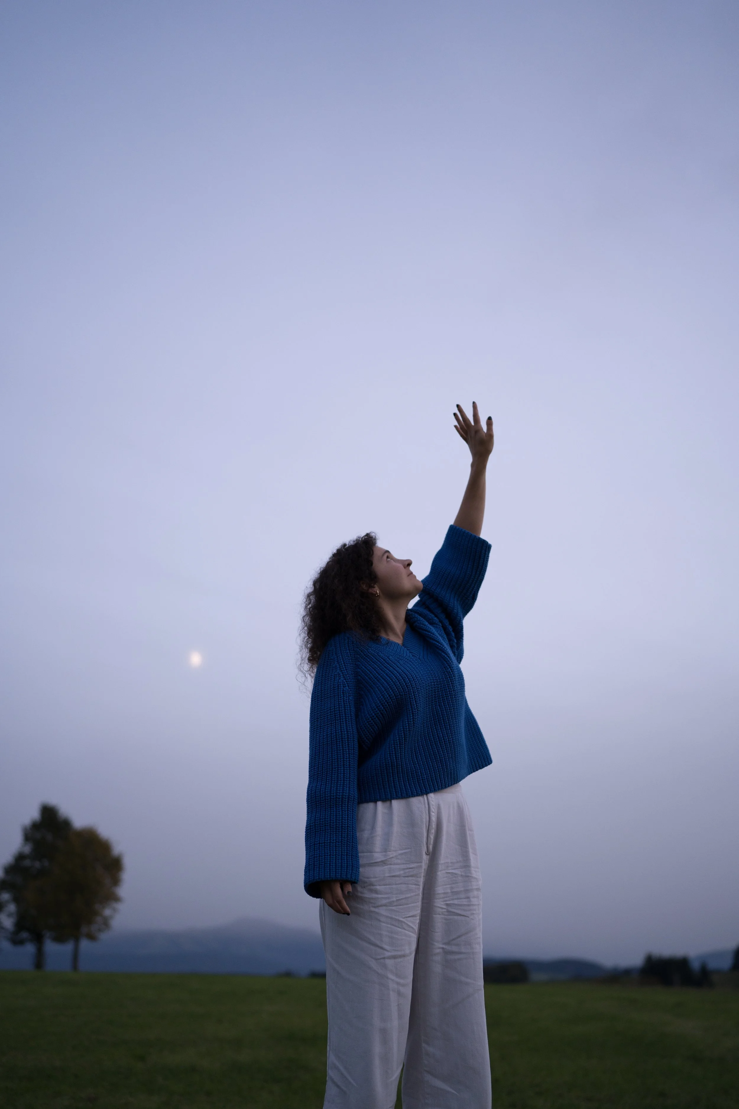 Eine junge Frau mit lockigem Haar in blauer Strickjacke und weißen Hosen streckt einen Arm hoch in den Himmel, während sie im Freien bei Dämmerung steht. Der Himmel ist grau-blau, mit sichtbarem Mond im Hintergrund, und es gibt Bäume und eine grüne Wiese im Hintergrund.