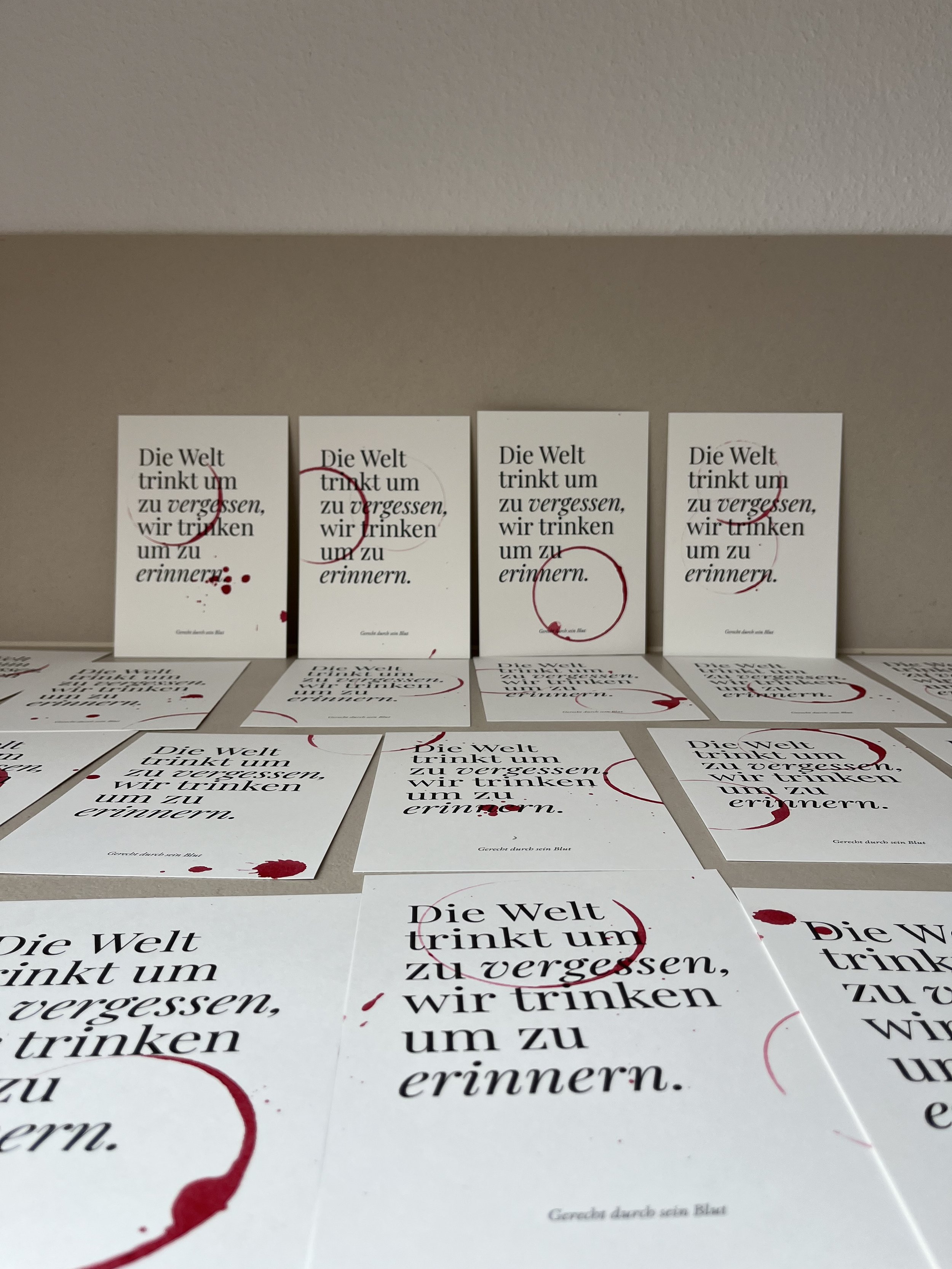 Mehrere weiße Karten mit schwarzem Text und roten Akzenten, die den Satz 'Die Welt trinkt um zu vergessen, wir trinken um zu erinnern.' enthalten, einige Karten stehen aufrecht an einer Wand, andere liegen flach auf einer Oberfläche.