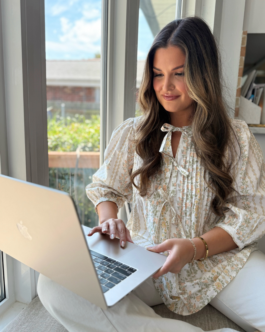 Nicole Merrell on Laptop Computer sitting by window in a flower blouse