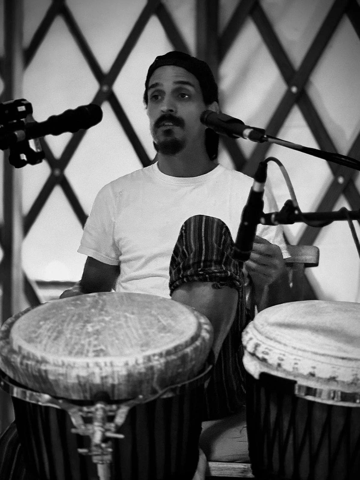 A man with dark hair, a beard, and mustache playing drums, surrounded by microphones, in a setting with a lattice background.