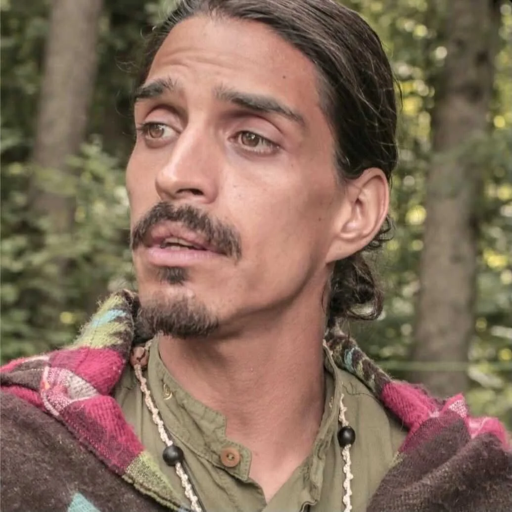 A man with medium-length dark hair, a mustache and goatee, looking to his left in an outdoor wooded setting.
