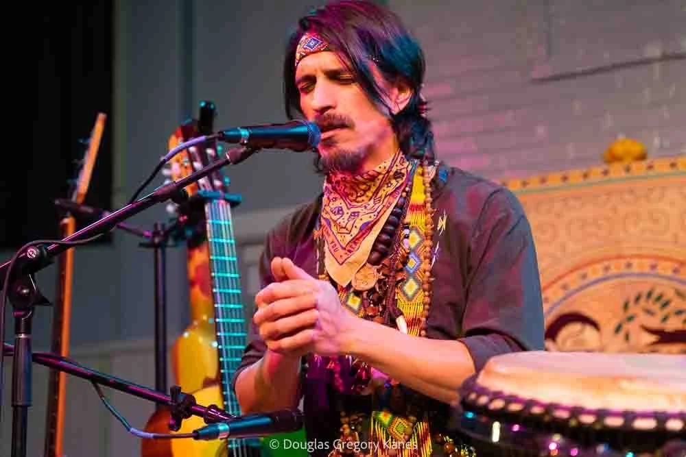 A musician with colorful hair and layered necklaces, singing into a microphone with a guitar in the background, performing on stage.