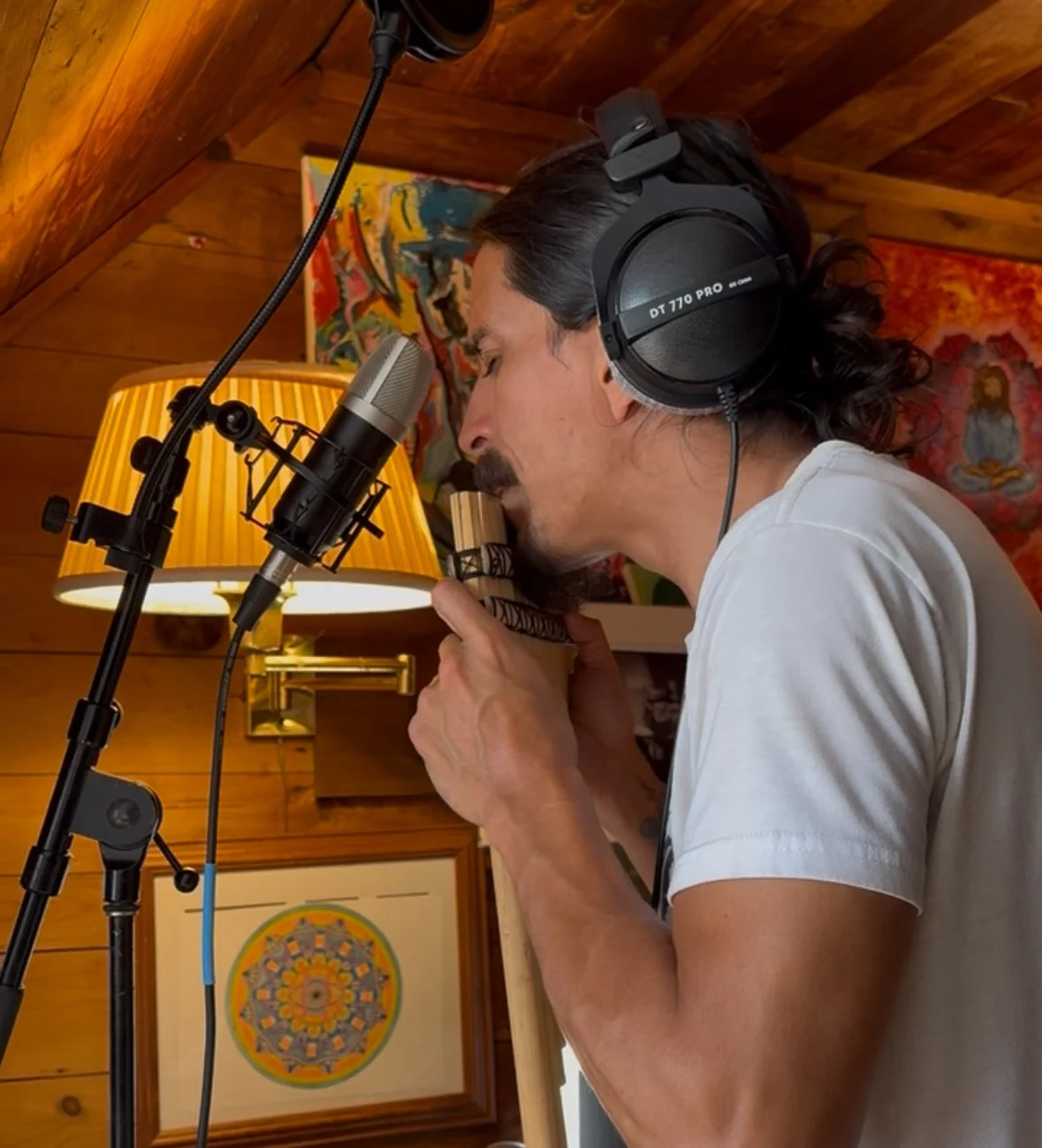 A man in a white t-shirt singing into a microphone in a cozy, wood-paneled room with colorful artwork and a yellow lamp in the background.