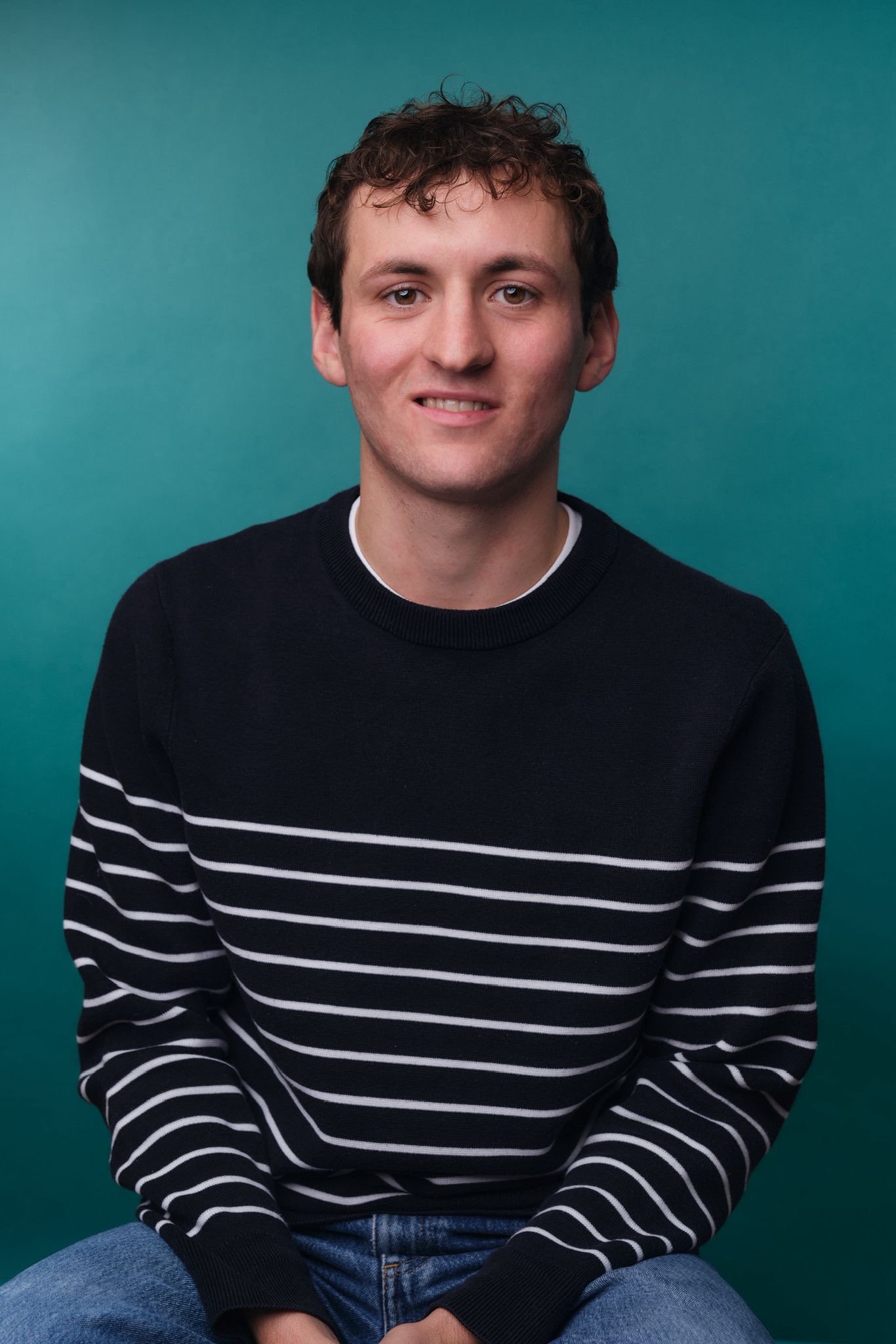 Portrait of a young man with curly brown hair, blue eyes, wearing a black and white striped sweater and jeans, sitting against a teal background.