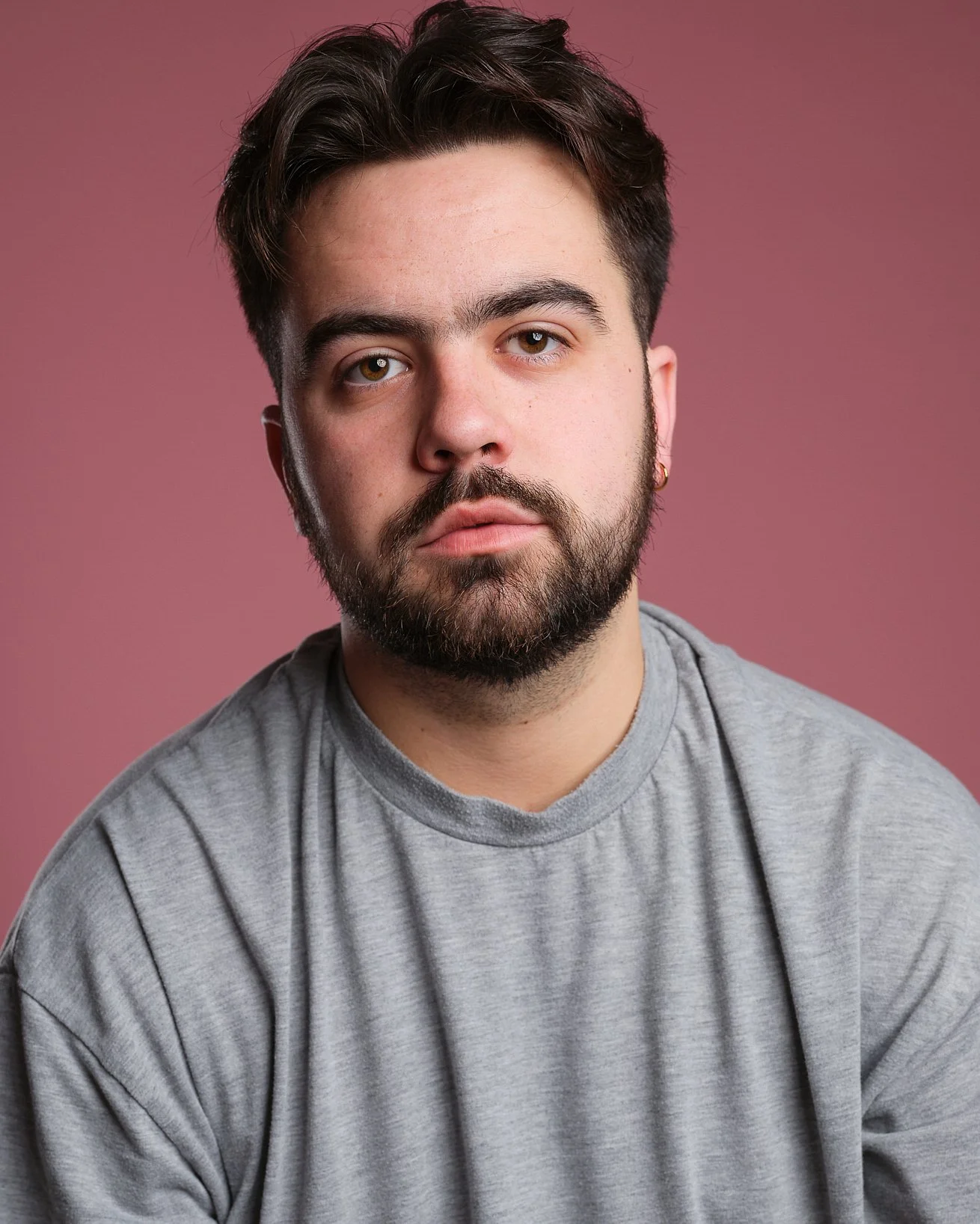 A young man with brown hair and a beard wearing a gray t-shirt, sitting against a pink background.
