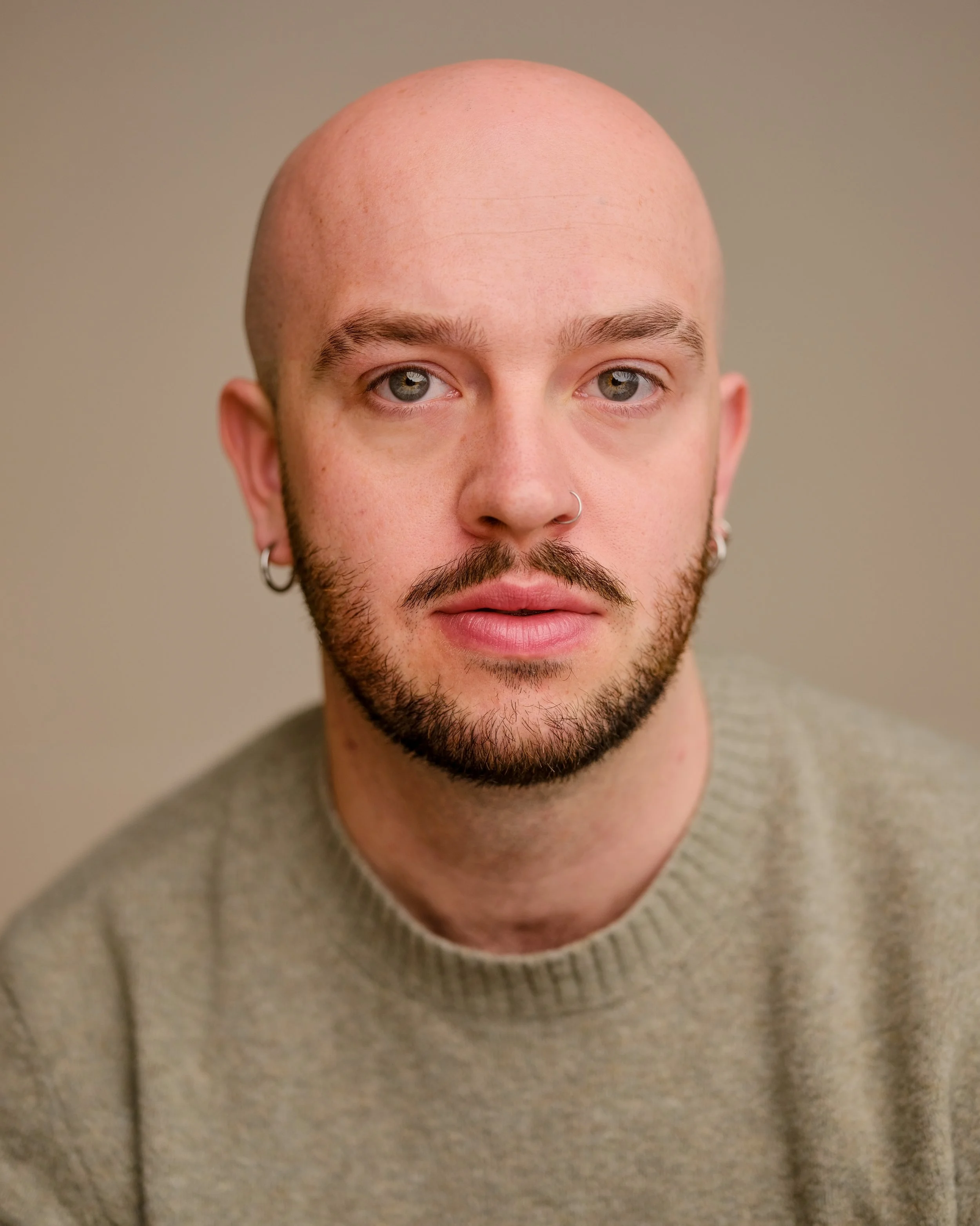 Close-up of a man with a shaved bald head, light skin, blue eyes, a beard, and multiple earrings in ears, wearing a beige sweater.