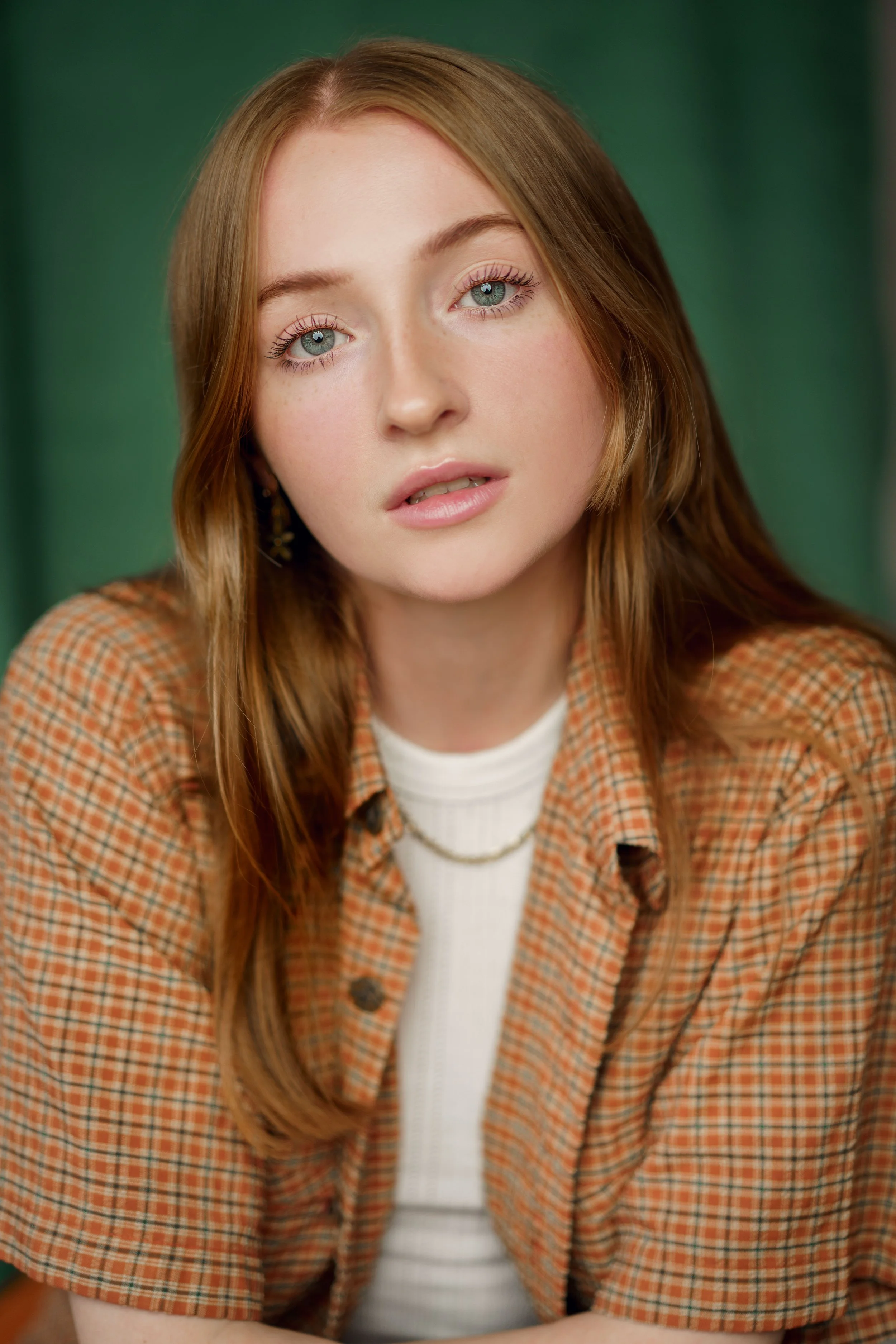 A young woman with red hair and blue eyes wearing a beige checked shirt over a white top, with a gold chain necklace, looking directly at the camera with a neutral expression.