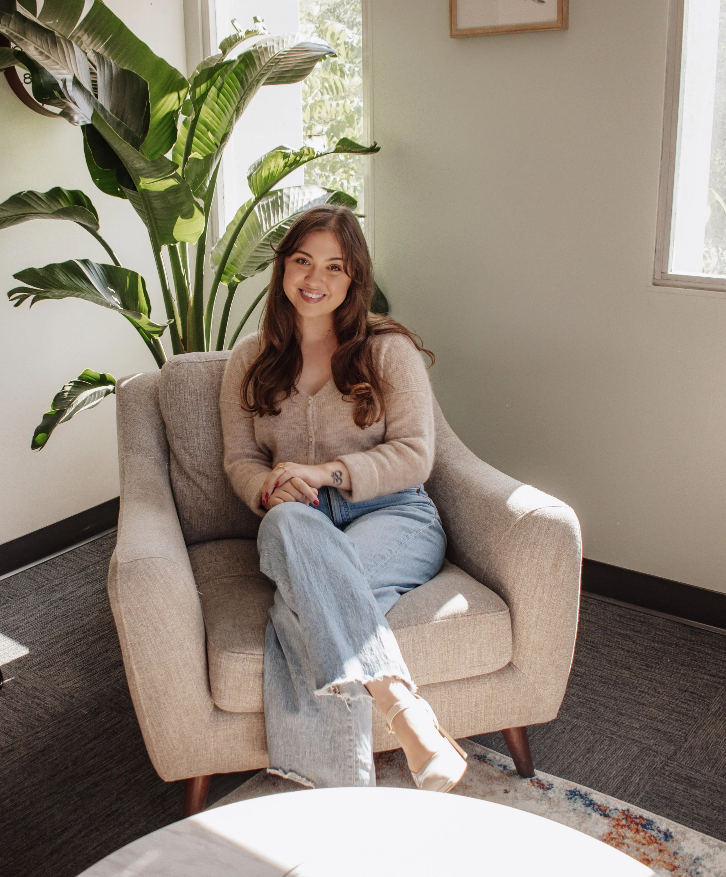 A woman with long brown hair, wearing a beige cardigan and ripped jeans, sitting on a beige armchair in a bright room, smiling at the camera. There is a large green leafy plant behind her and a window through which natural light enters.