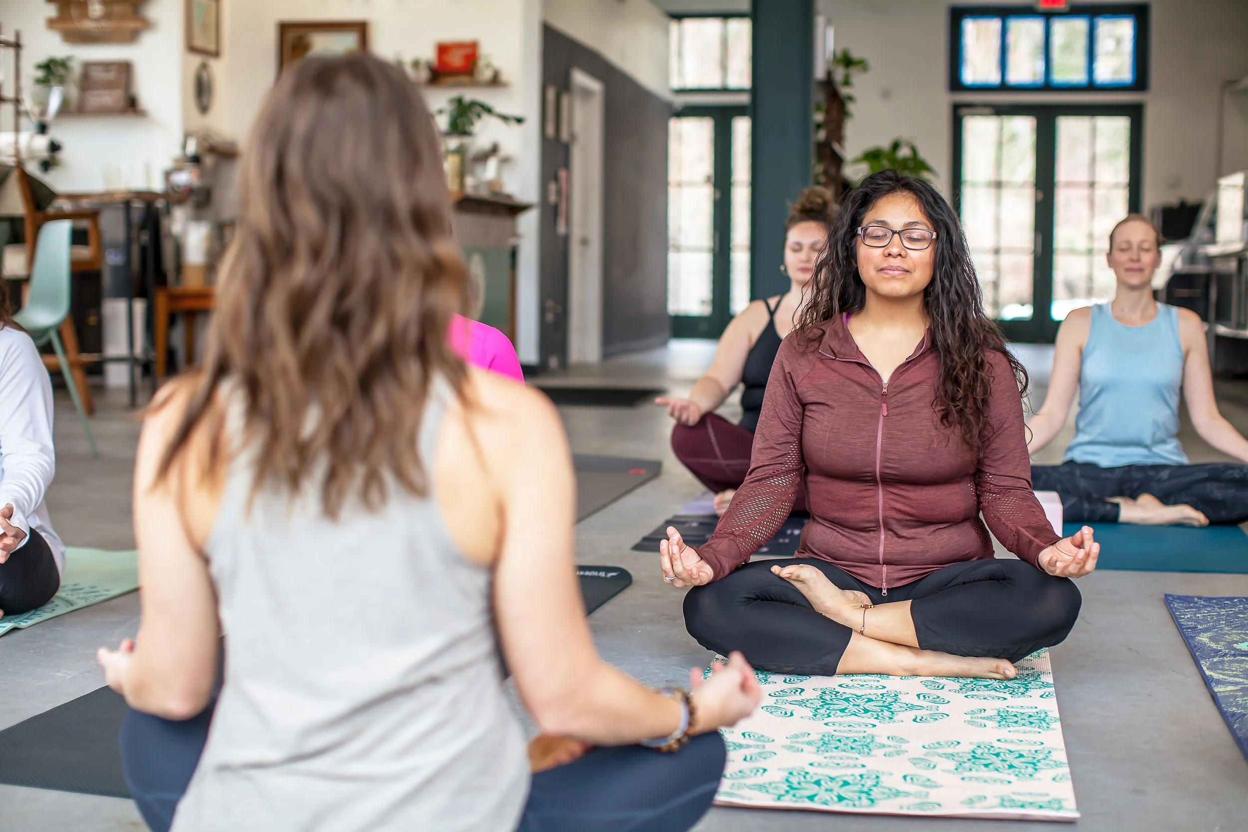 Group yoga meditation class with women sitting cross-legged on mats inside a studio.