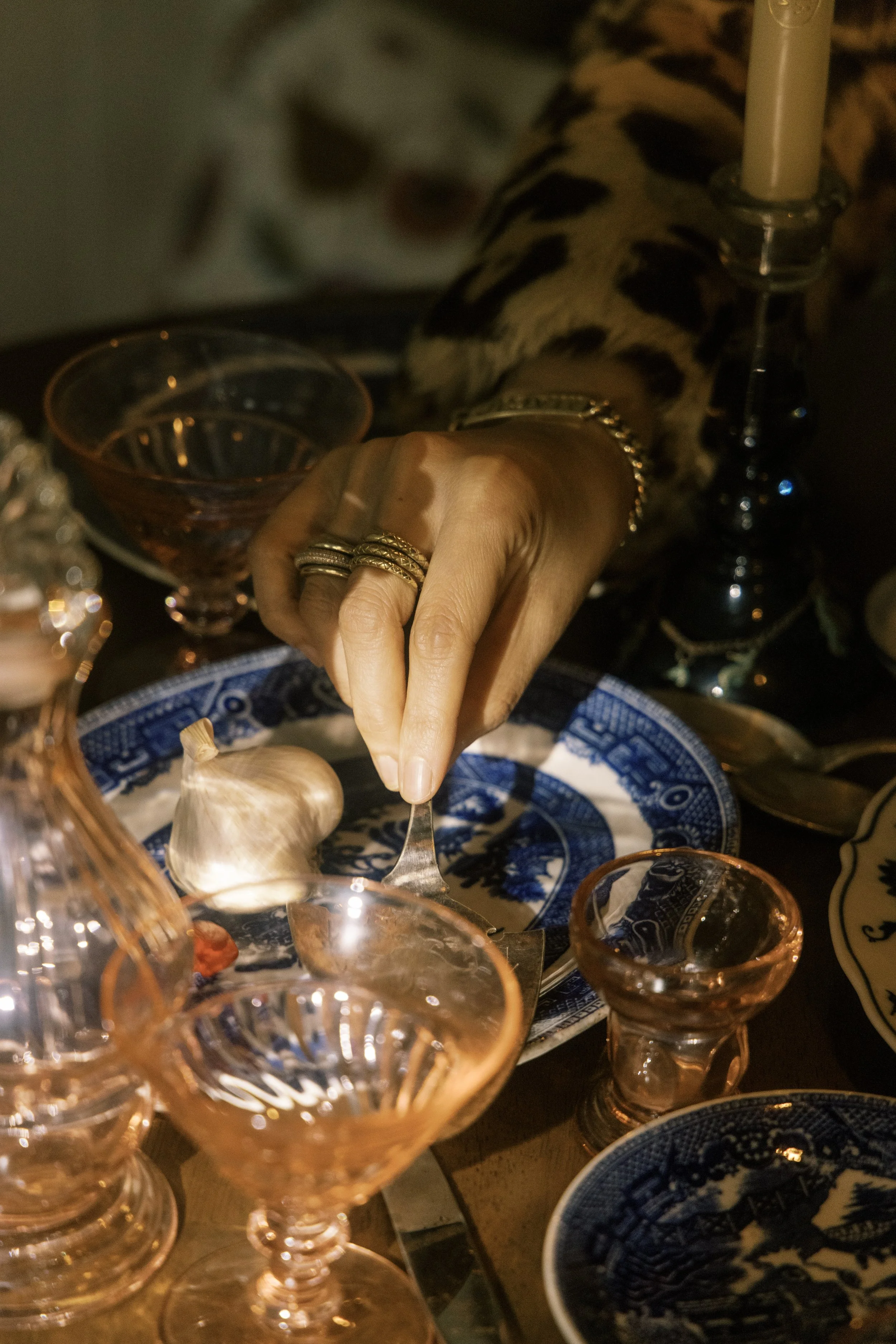 A person reaching for food with a fork at a table set with vintage-style glassware, patterned blue and white plates, and a large piece of garlic. The person is wearing multiple rings and a chain bracelet, and is dressed in a leopard print top. The scene is dimly lit, creating a warm, intimate atmosphere.