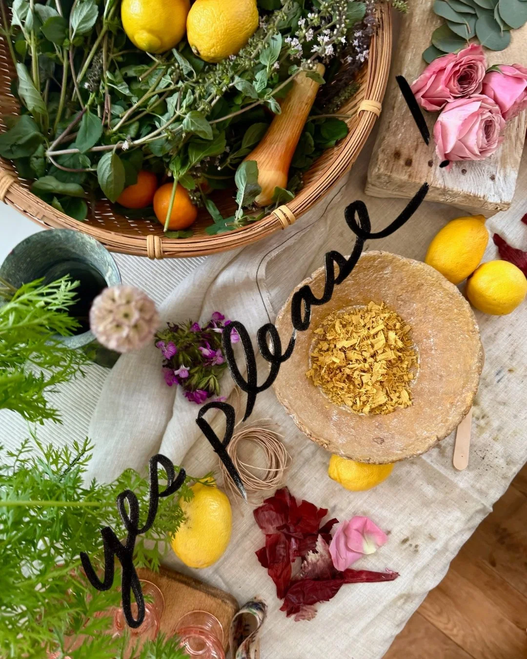 A rustic table setting with a basket of fresh lemons, oranges, and greenery, a small glass of dark liquid with a plant, a plate of crushed basil or turmeric, pink roses, purple and red flowers, and a handwritten sign that says 'Pepper & Lace'.