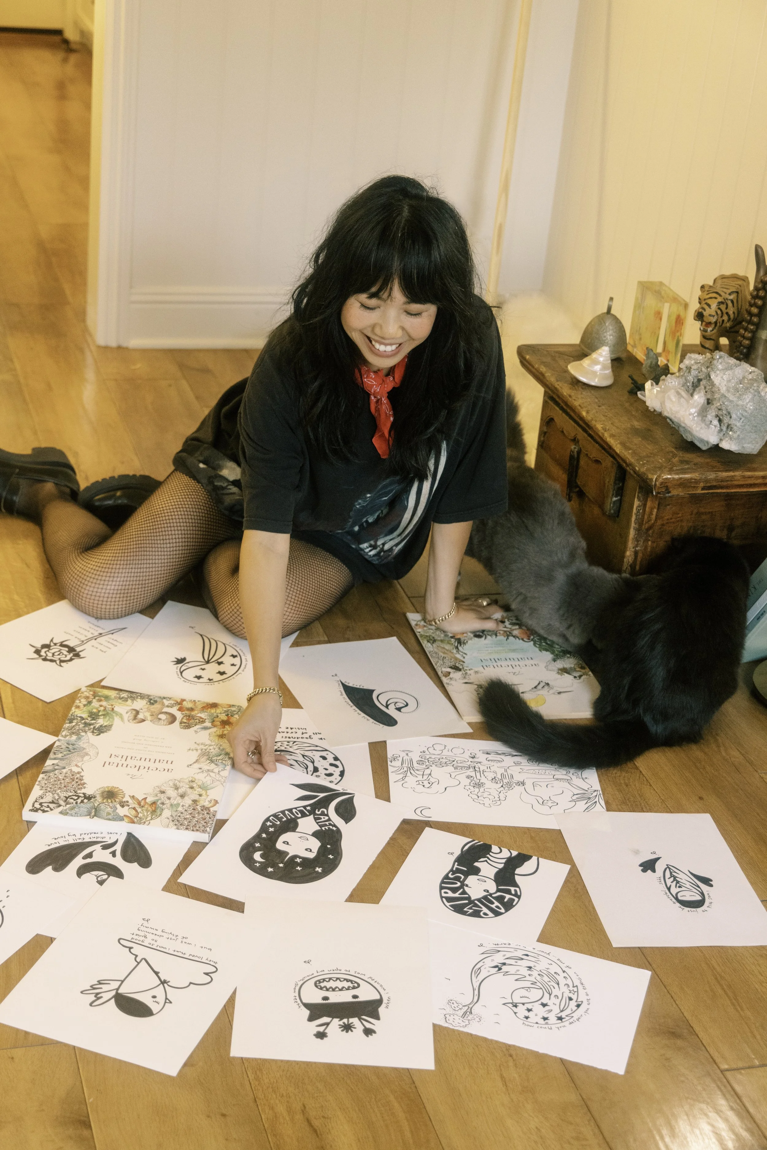 Woman with black hair, black t-shirt, fishnet stockings, and black boots, sitting on wooden floor surrounded by black-and-white artwork, with a black cat nearby.