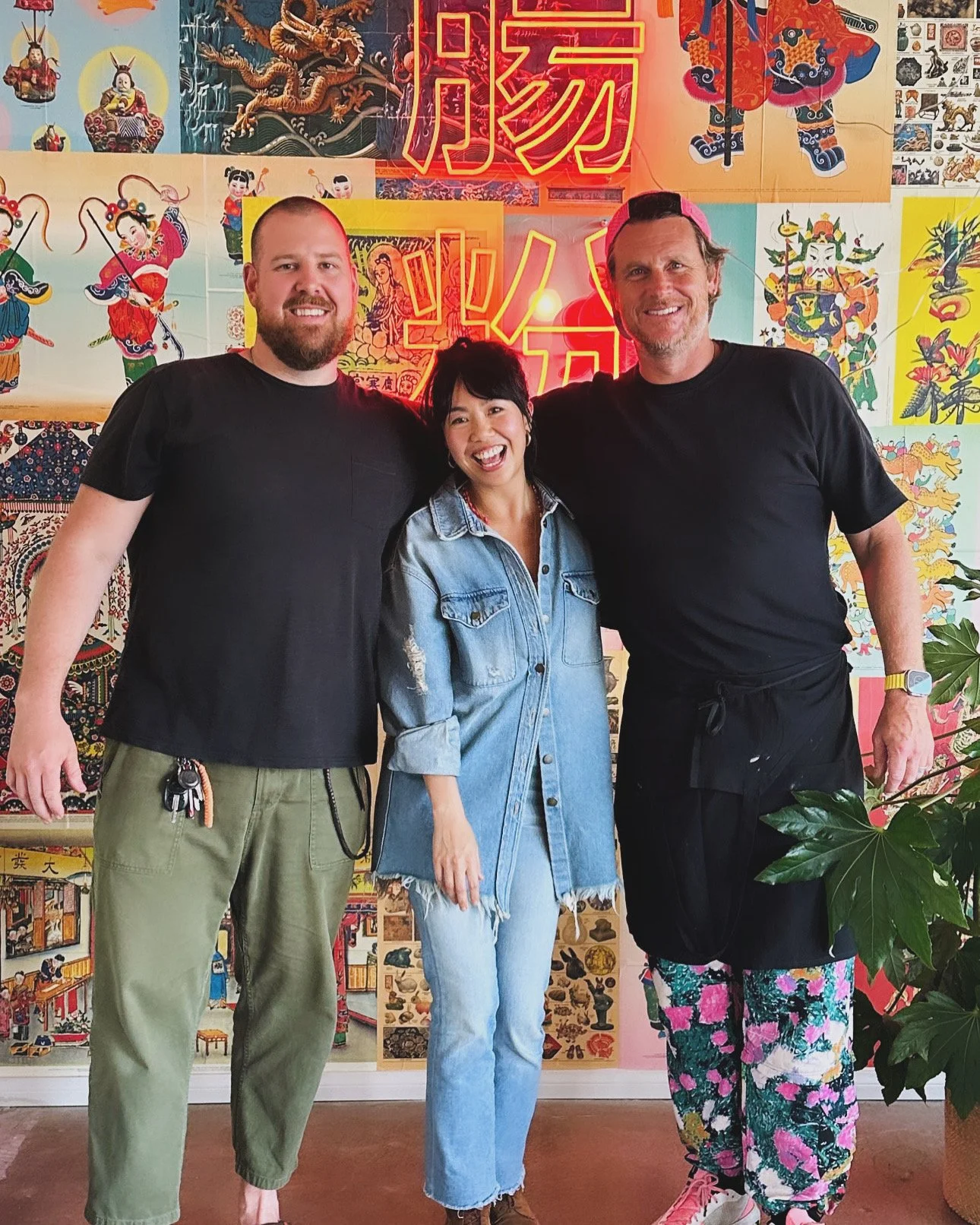 Three people standing together in front of a colorful wall decorated with Asian art and neon red Chinese characters. The group includes two men and one woman, all smiling. The man on the left has a beard, is wearing a black T-shirt and green pants. The woman in the middle has black hair, is wearing a light blue denim jacket and jeans. The man on the right has gray hair, is wearing a black T-shirt, floral pants, and a pink cap.