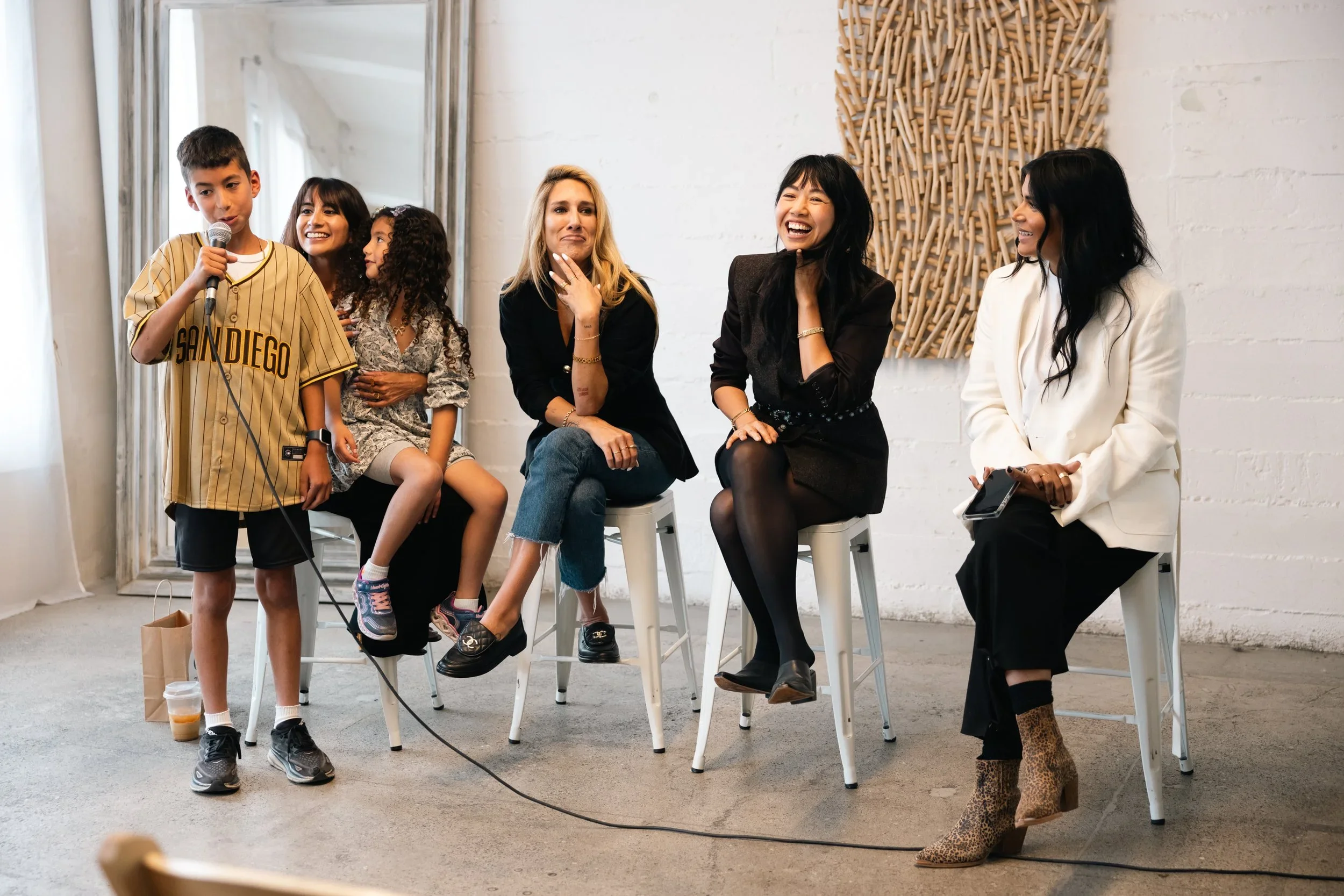 Group of six diverse people, five women and one boy, sitting on bar stools and one boy standing, laughing and talking in a bright room with white brick walls and large mirrors.